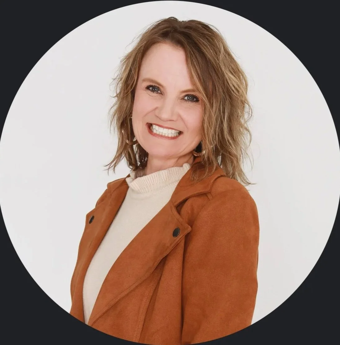 A woman with shoulder-length blond hair smiling, wearing a beige sweater and a brown jacket, against a white background.