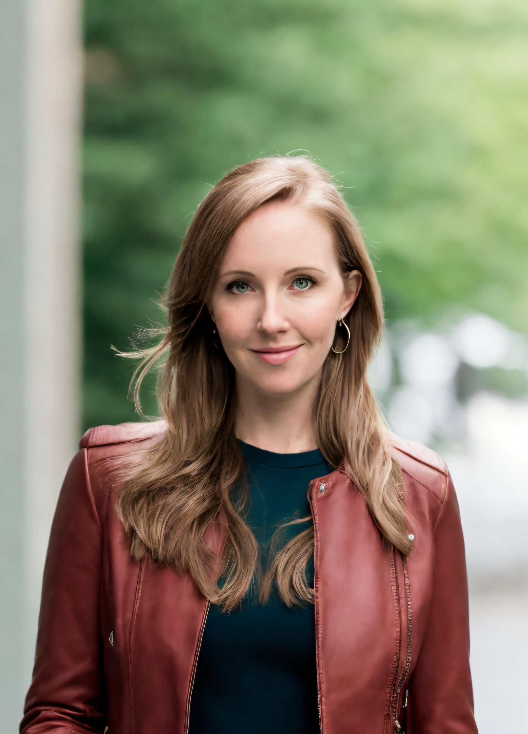 A woman with long wavy light brown hair, wearing a red leather jacket and a dark blue shirt, smiling outdoors with green blurred background.