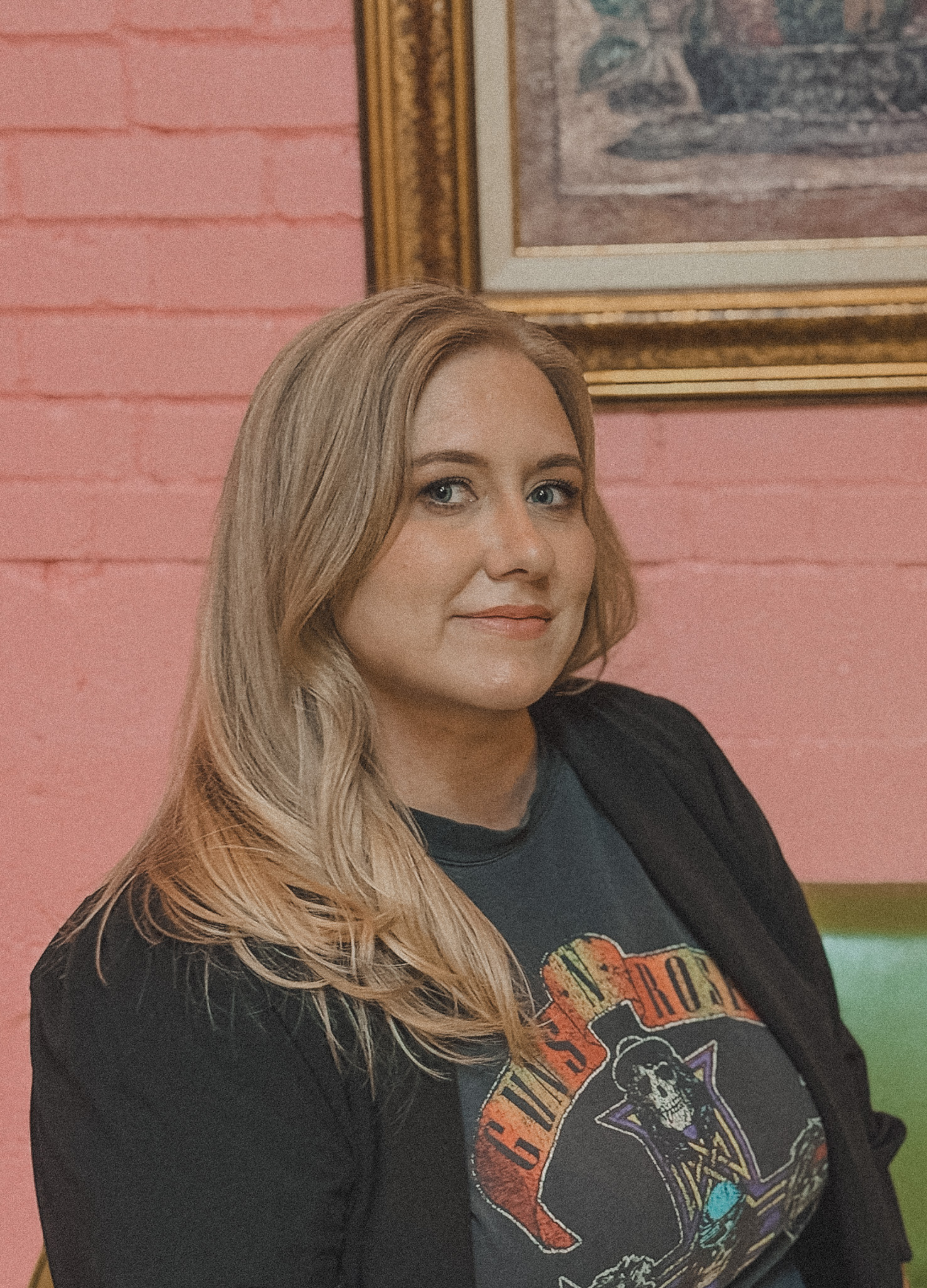 A woman with long blonde hair wearing a graphic Guns N' Roses t-shirt and black jacket, sitting against a pink brick wall background.