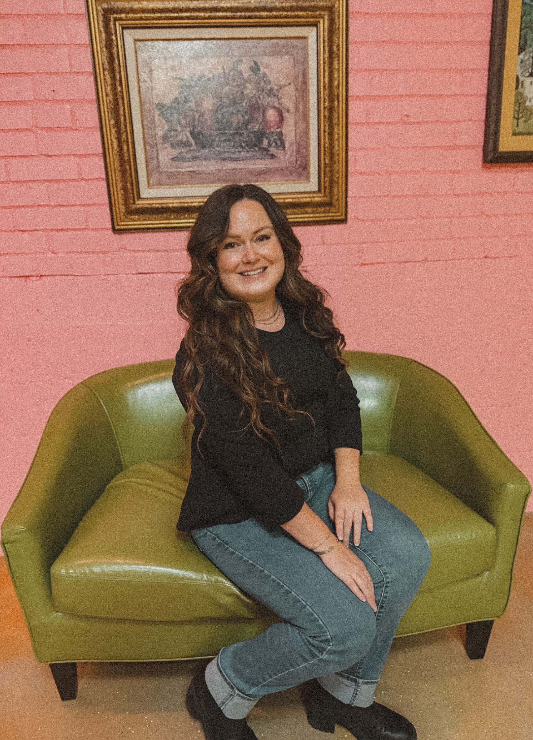 A woman with long, wavy brown hair smiling and sitting on a green couch in front of a pink brick wall with framed artwork.