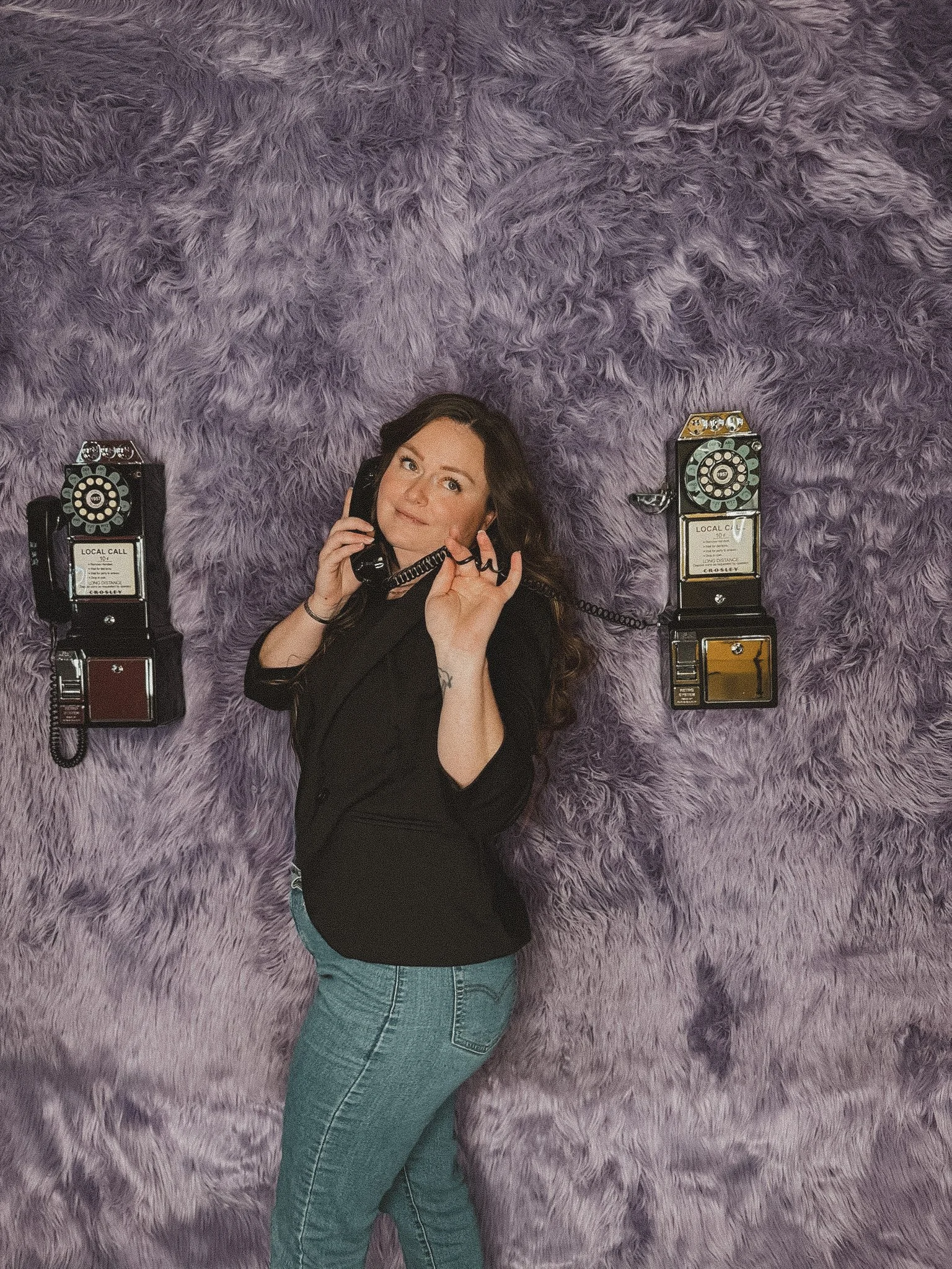 Woman with long dark hair lying on a purple fluffy carpet, holding a vintage black rotary telephone to her ear and making an 'okay' gesture with her hand.