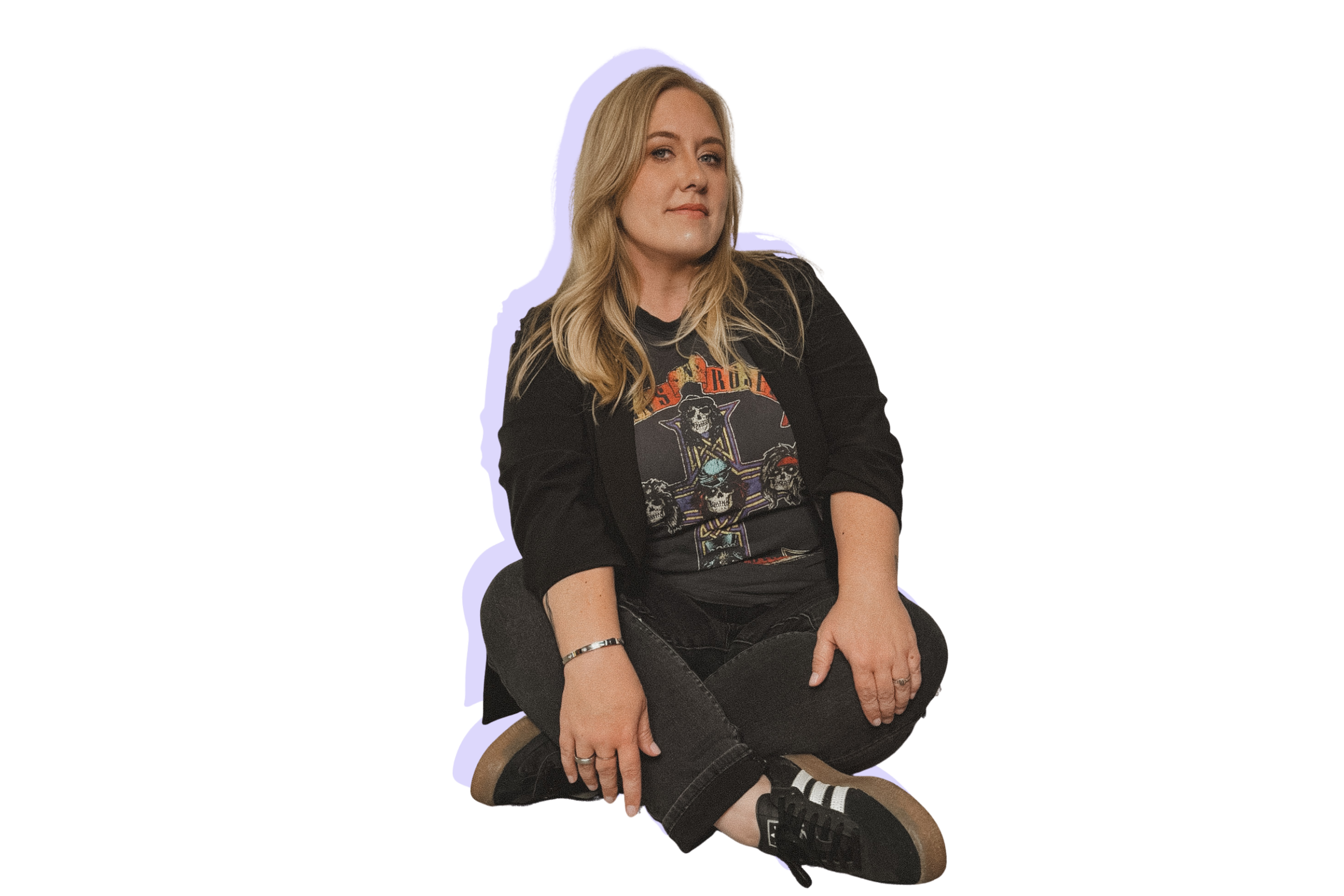 A woman with blonde hair sitting cross-legged on the floor, wearing a black vintage rock t-shirt, black jeans, and black sneakers with white stripes.