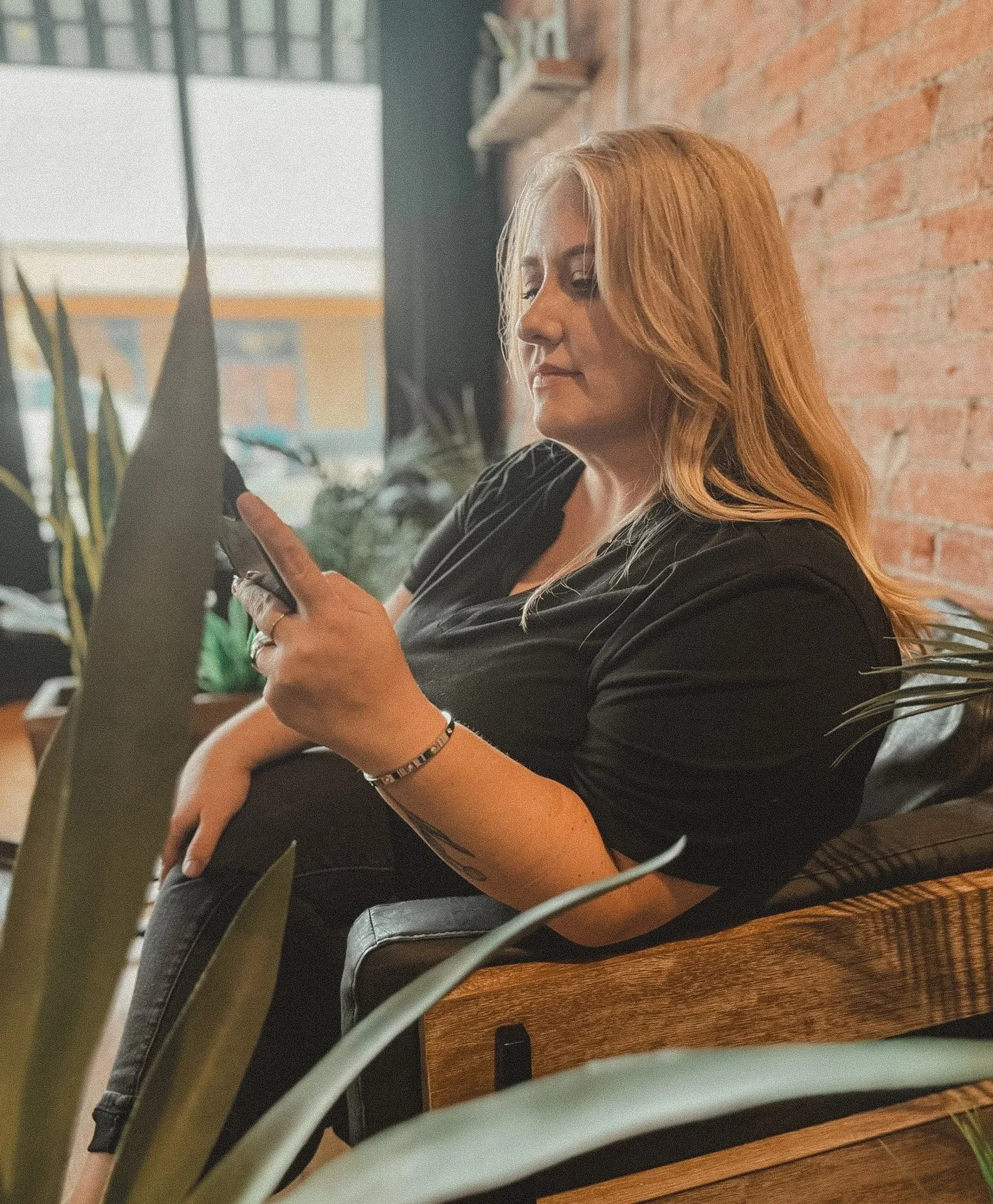 Woman with long red hair sitting on a wooden bench, looking at her phone, in a cozy indoor setting with exposed brick wall and plants.