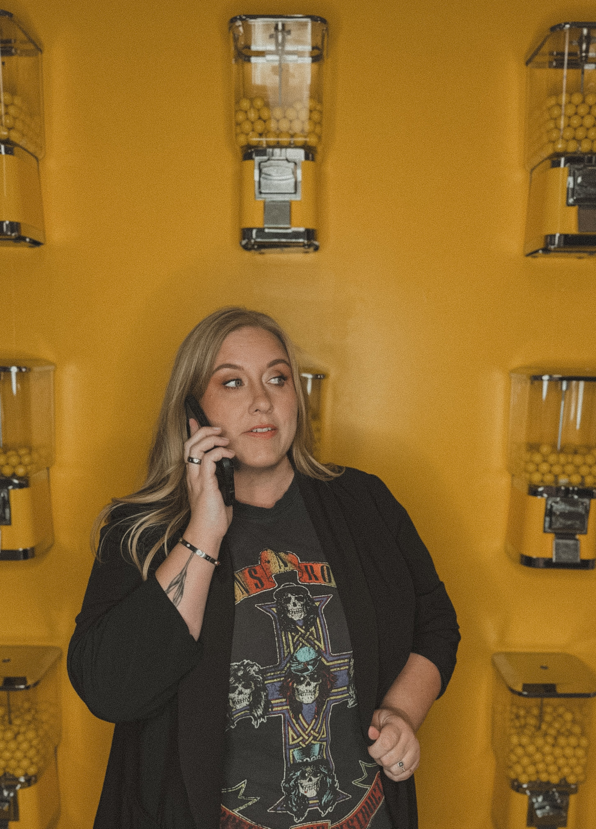 A woman talking on a cellphone in front of yellow gumball vending machines, wearing a black jacket and a graphic t-shirt.