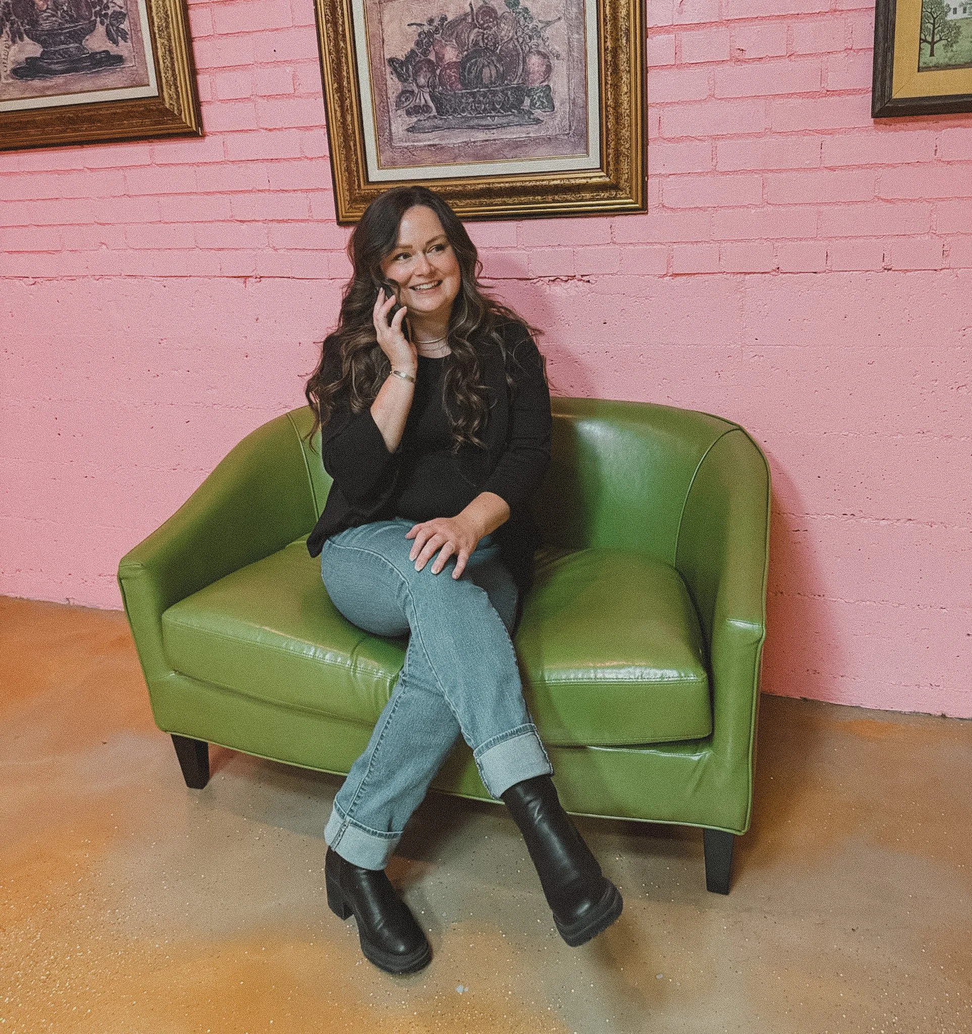 A woman with long dark curly hair sitting on a bright green sofa, smiling and talking on a cellphone, with a pink brick wall and framed artwork behind her.
