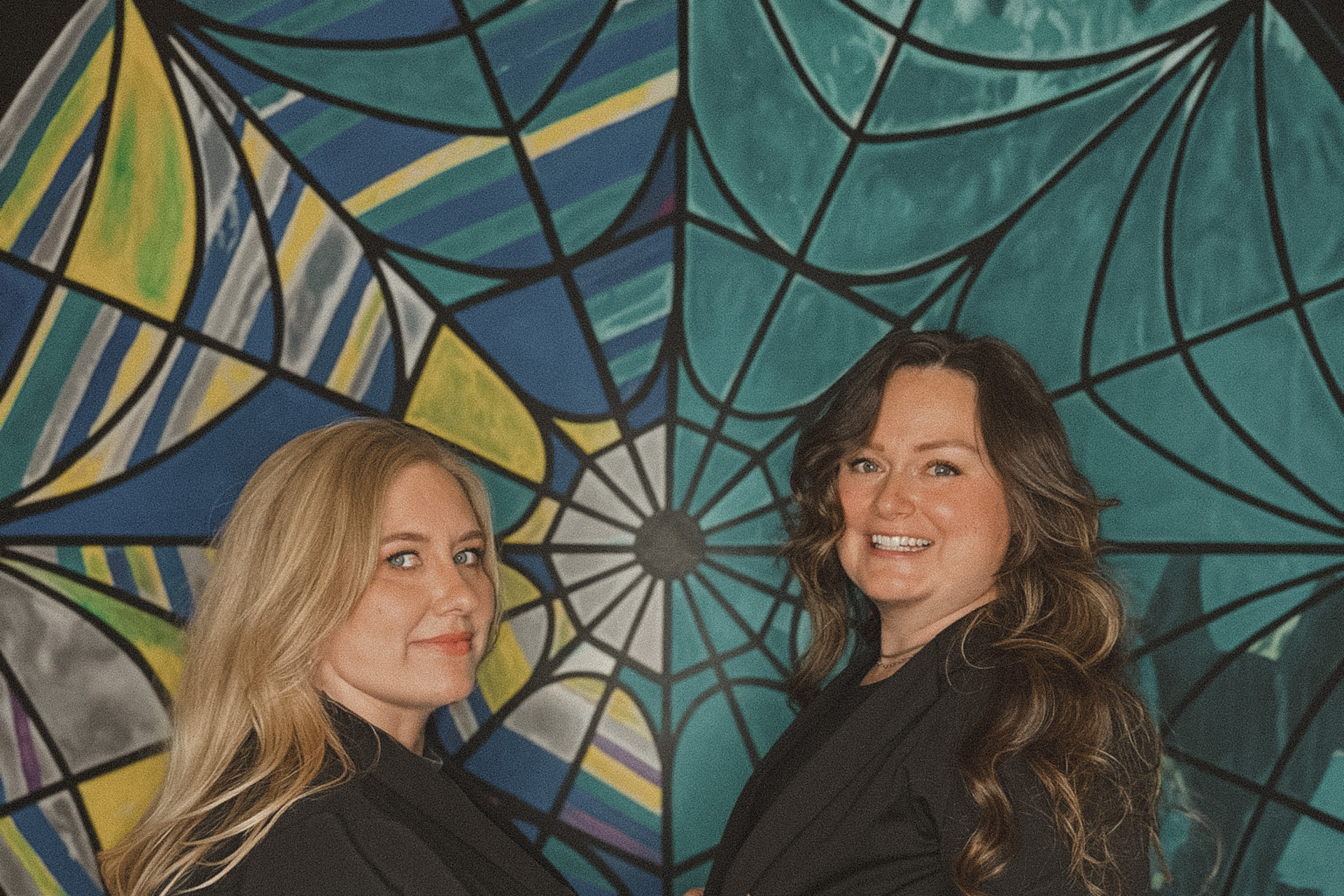 Two women standing in front of a large, colorful mural featuring a spiderweb design in shades of blue, green, yellow, gray, and purple.