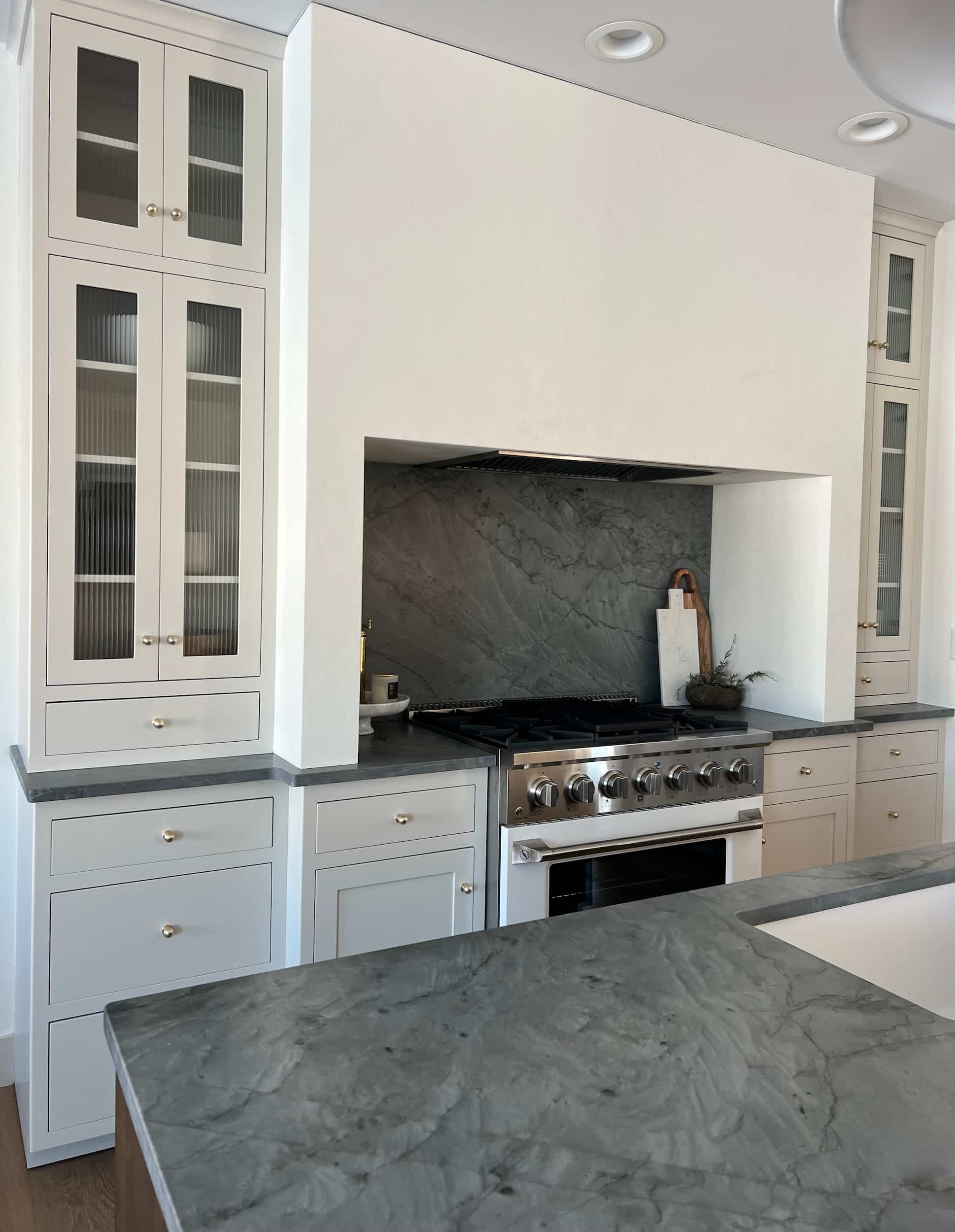 View across the dark kitchen island counter toward the range and soapstone backsplash. Tall white cabinets with glass inserts flank the range hood area.