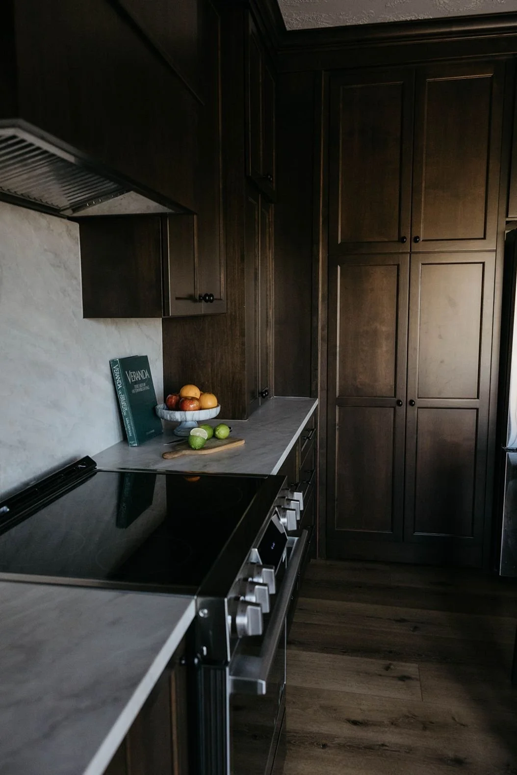 Dark wood kitchen cabinetry surrounding a modern stainless steel range and a marble slab backsplash. The countertop is styled with a bowl of fruit and a cookbook.