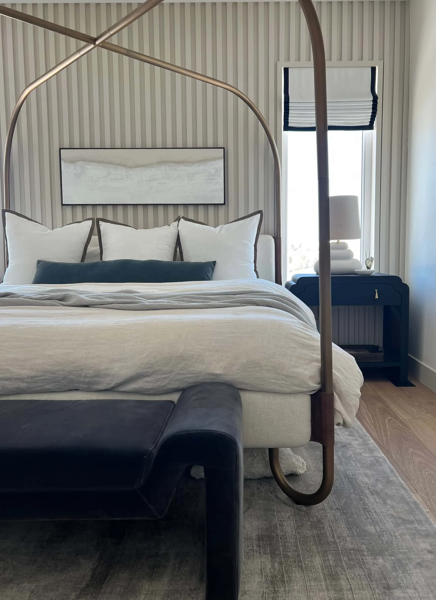 Master bedroom full view showcasing a curved brass four-poster canopy bed, layered neutral bedding, a dark velvet bench at the foot, and a ribbed/slatted accent wall.