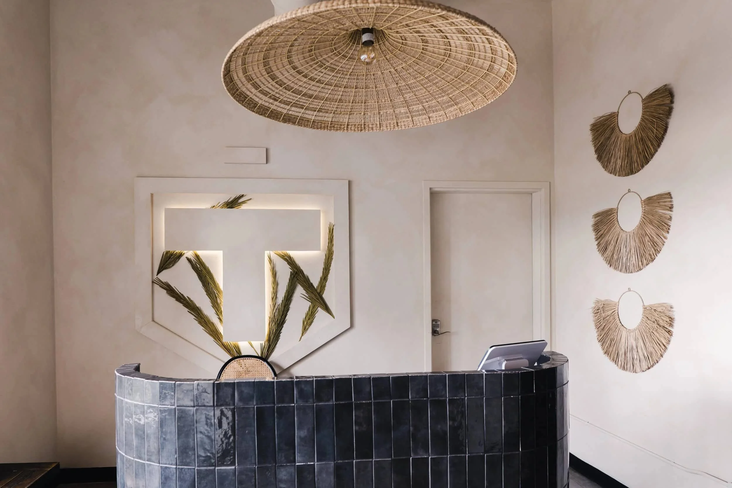 Overview of the reception area, featuring the black tiled desk, 'T' logo wall art, rattan chandelier, and a decorative wall hanging.