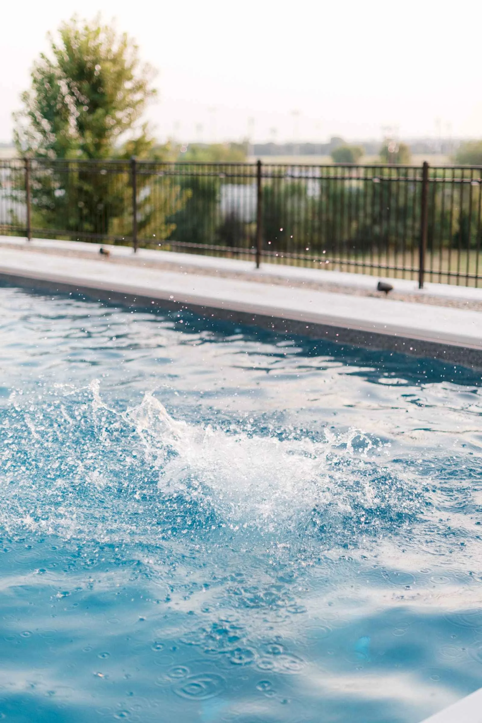 Close-up of clear blue pool water with a large splash near the edge, showing the white coping and black fence.