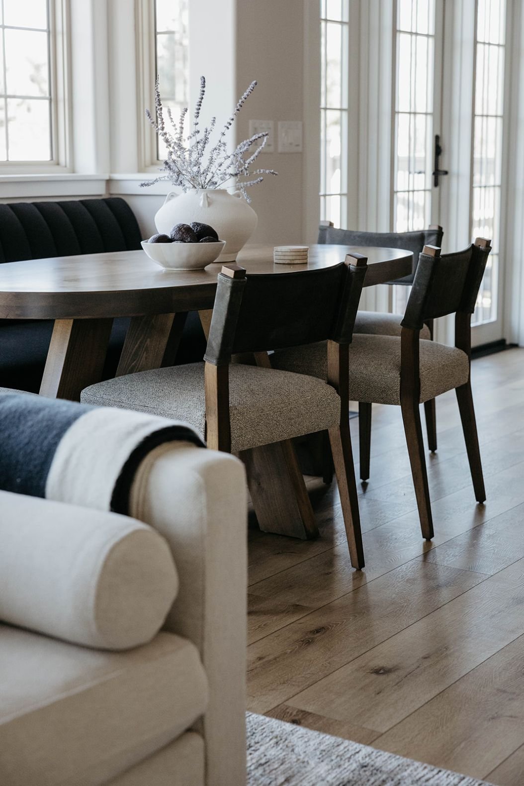 Close-up of the round wood dining table styled with a tall white vase. The shot is taken over the neutral sectional sofa in the adjacent living room