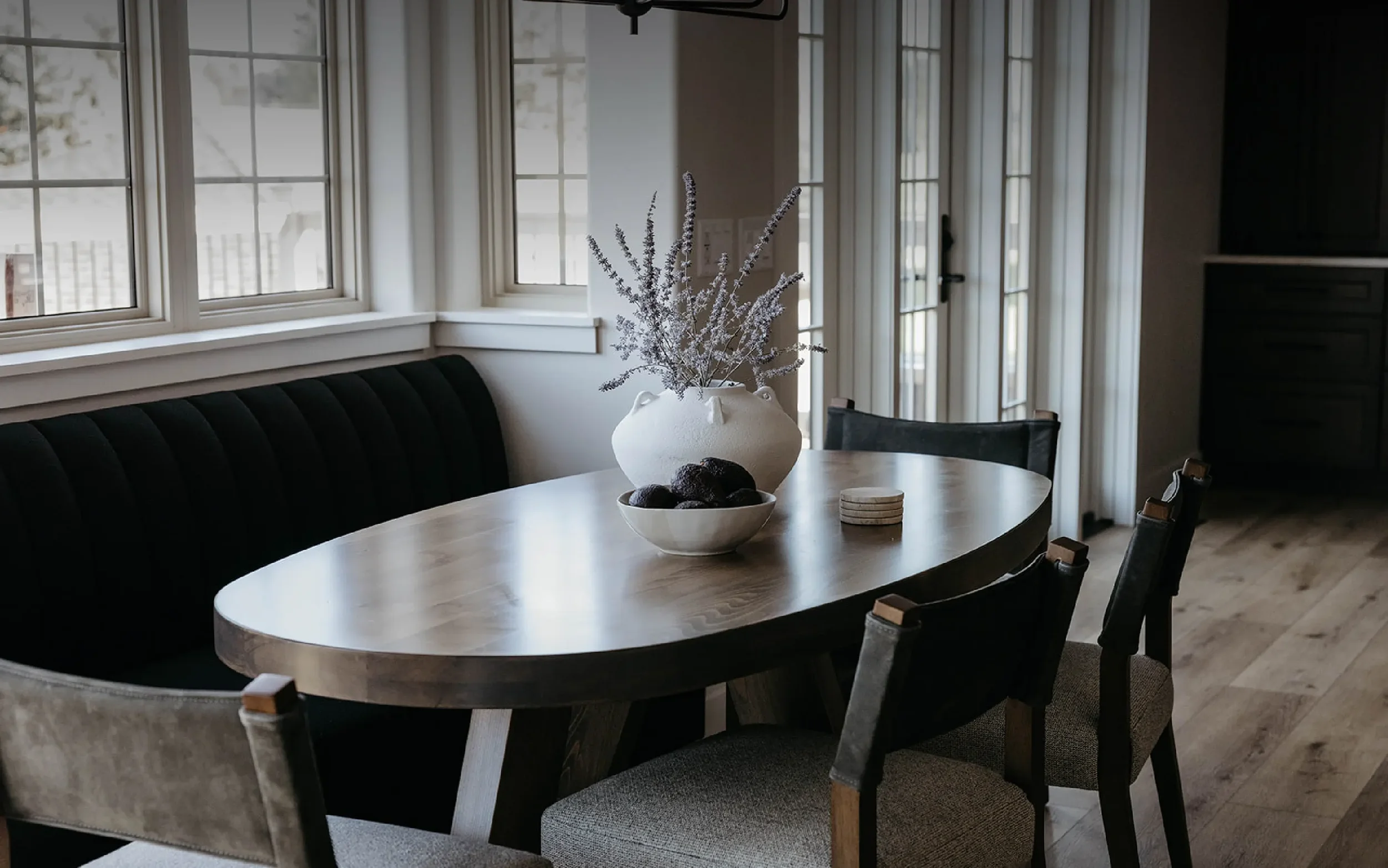 A dining room with a wooden oval table, black cushioned bench, and four chairs. A white vase with lavender flowers and a bowl of dark decorative stones are on the table. Windows with white trim let in natural light, and light-colored curtains hang be