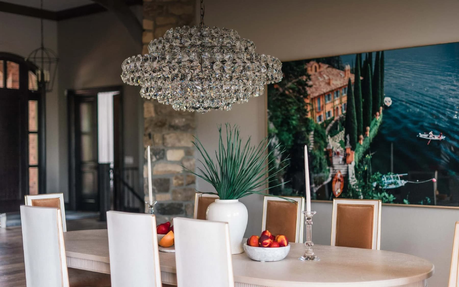 A dining room with a large crystal chandelier, a white table with six chairs, a vase with green leaves, two candlesticks, and a bowl of peaches. There is a large wall art piece showing a house and water scene in the background.