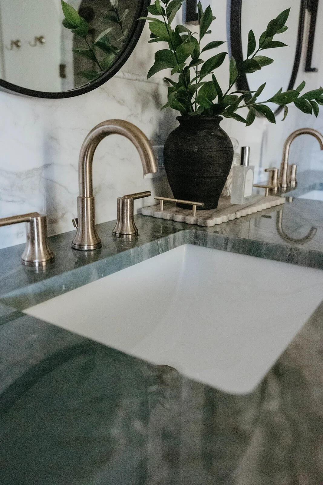 Close-up of a square bathroom sink set into a dark green marble countertop, featuring a brushed gold widespread faucet and a black vase with greenery.