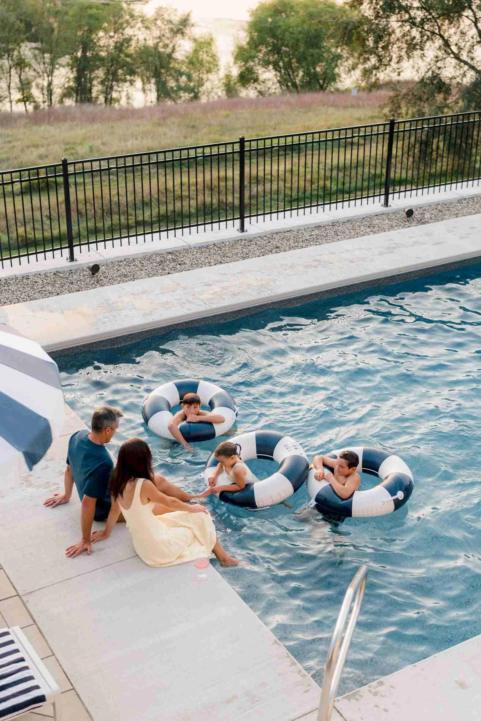 Overhead view of Christy Steen's family sitting on the pool ledge, wading in the water, and relaxing with striped floats on a sunny evening.