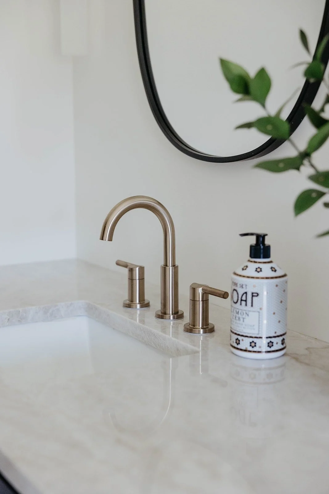 Close-up of a white marble countertop with subtle veining, showing a brushed gold widespread faucet and a decorative soap dispenser next to an under-mount sink.