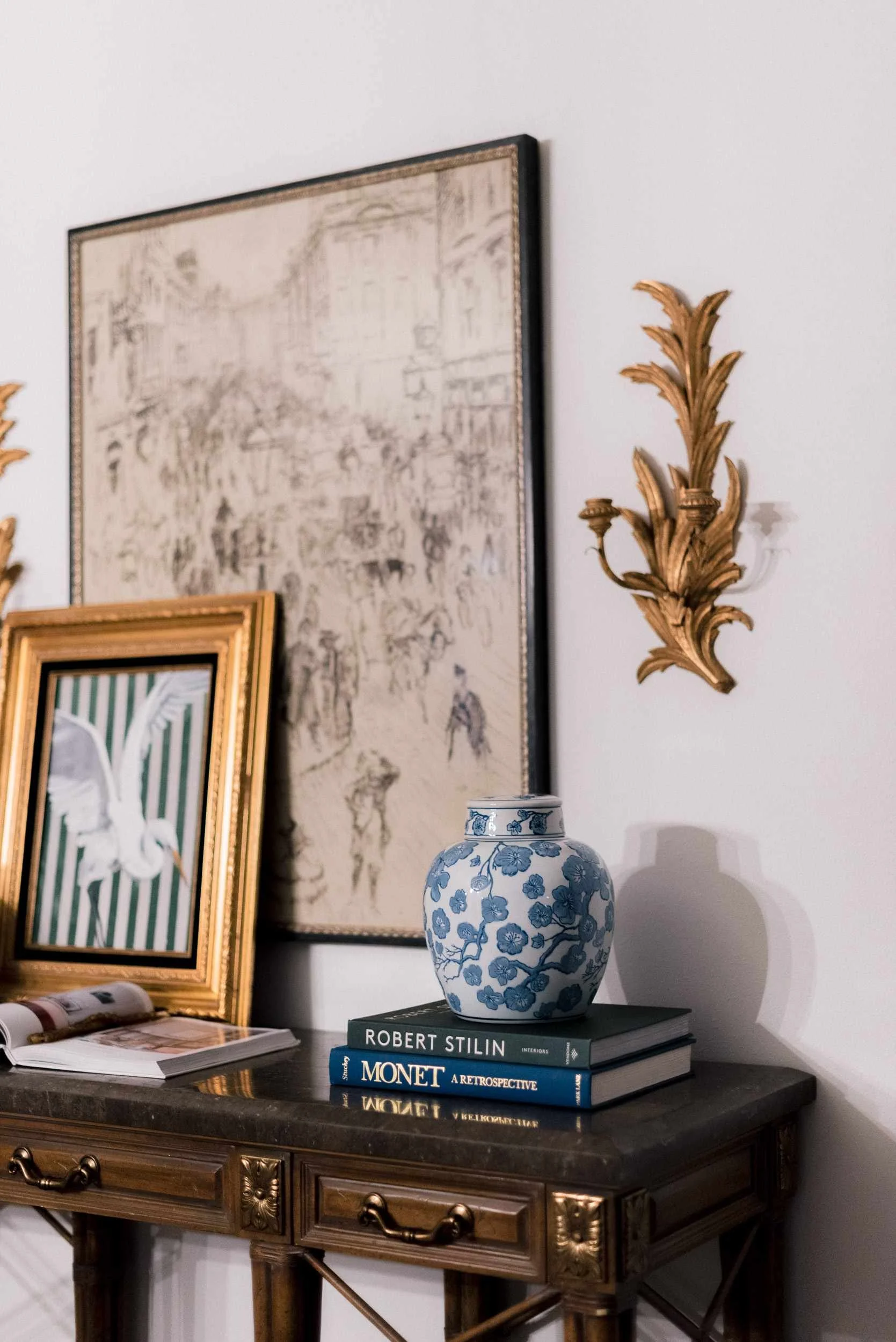 Detail view of the console table's marble surface, showing the blue and white ginger jar, an original painting by Christy Steen of a bird in a gold frame, and a large framed sketch of a busy scene.