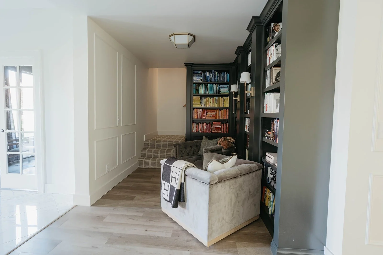View of a dark, custom-built library or office through a light hallway. The library features dark green or black built-in shelving and a light-colored accent chair.