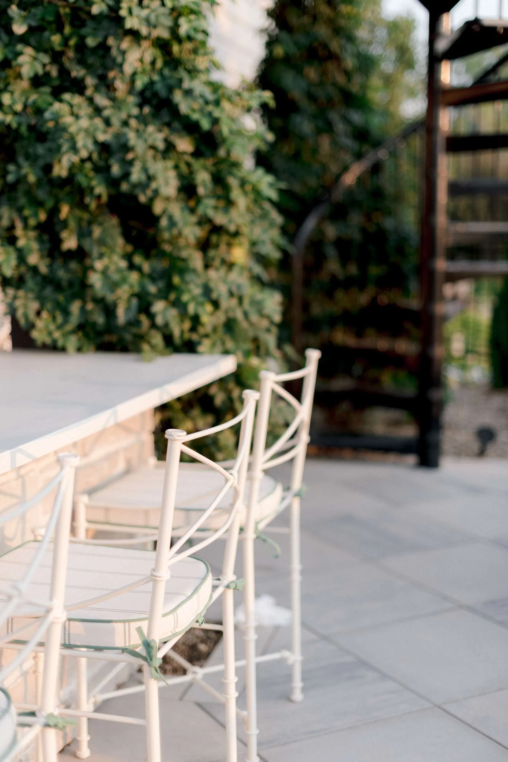 Close-up of white metal barstools with striped cushions tucked under a white outdoor bar counter, with a blurred view of the spiral staircase.