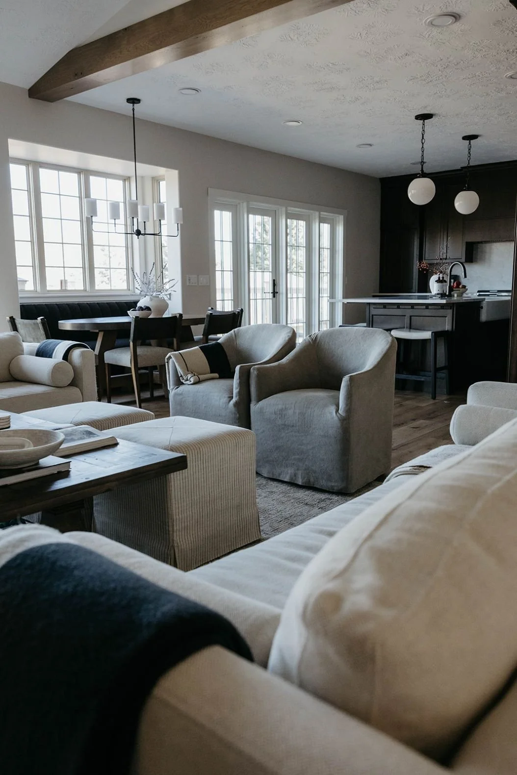 View of an open-concept living space with vaulted ceilings and exposed wood beams, looking past a sitting area toward the dining area and dark kitchen cabinetry.