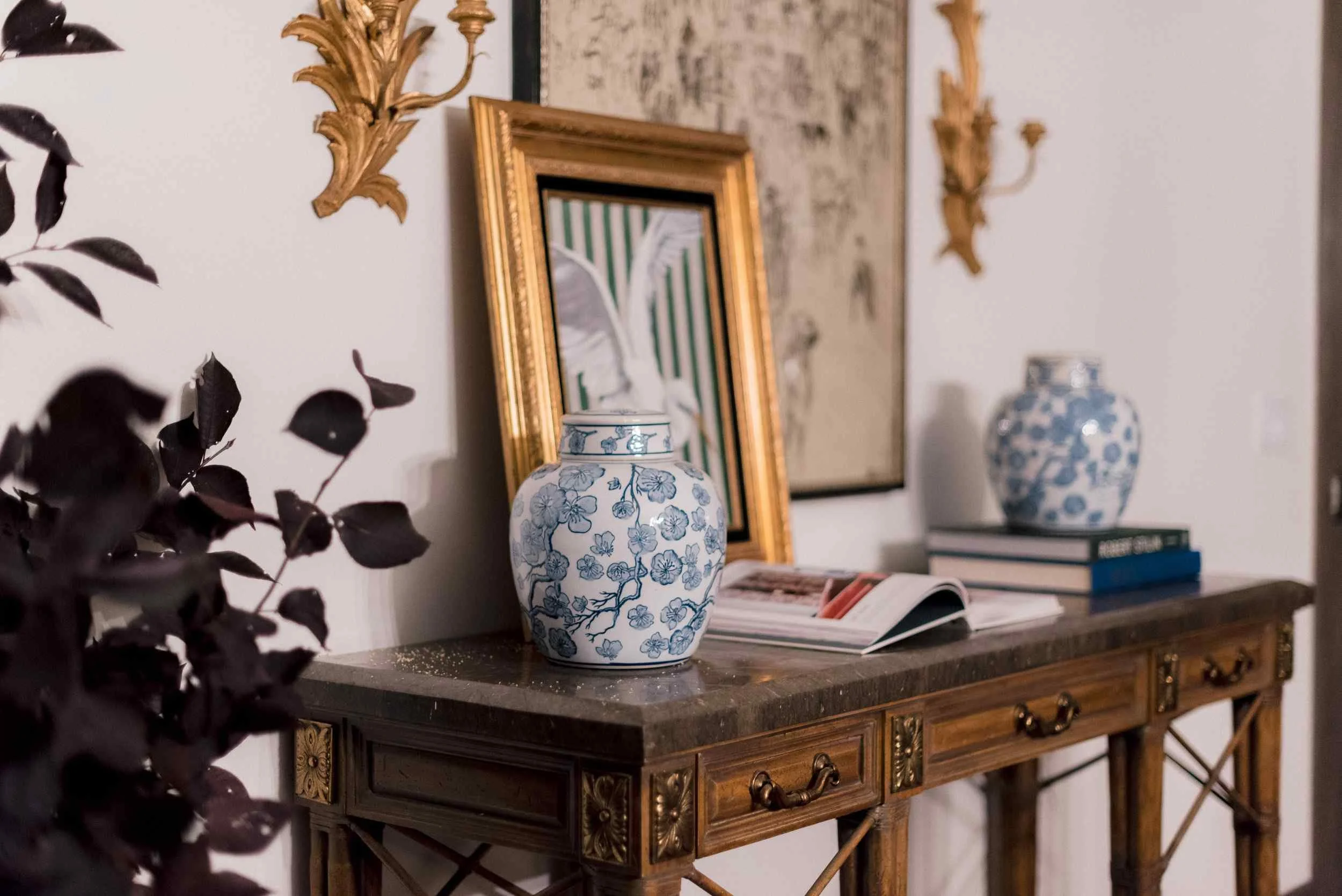 Close-up of the styled console table, showing the framed original artwork by Christy Steen, stacked blue books, and a gilt sconce on the white wall.