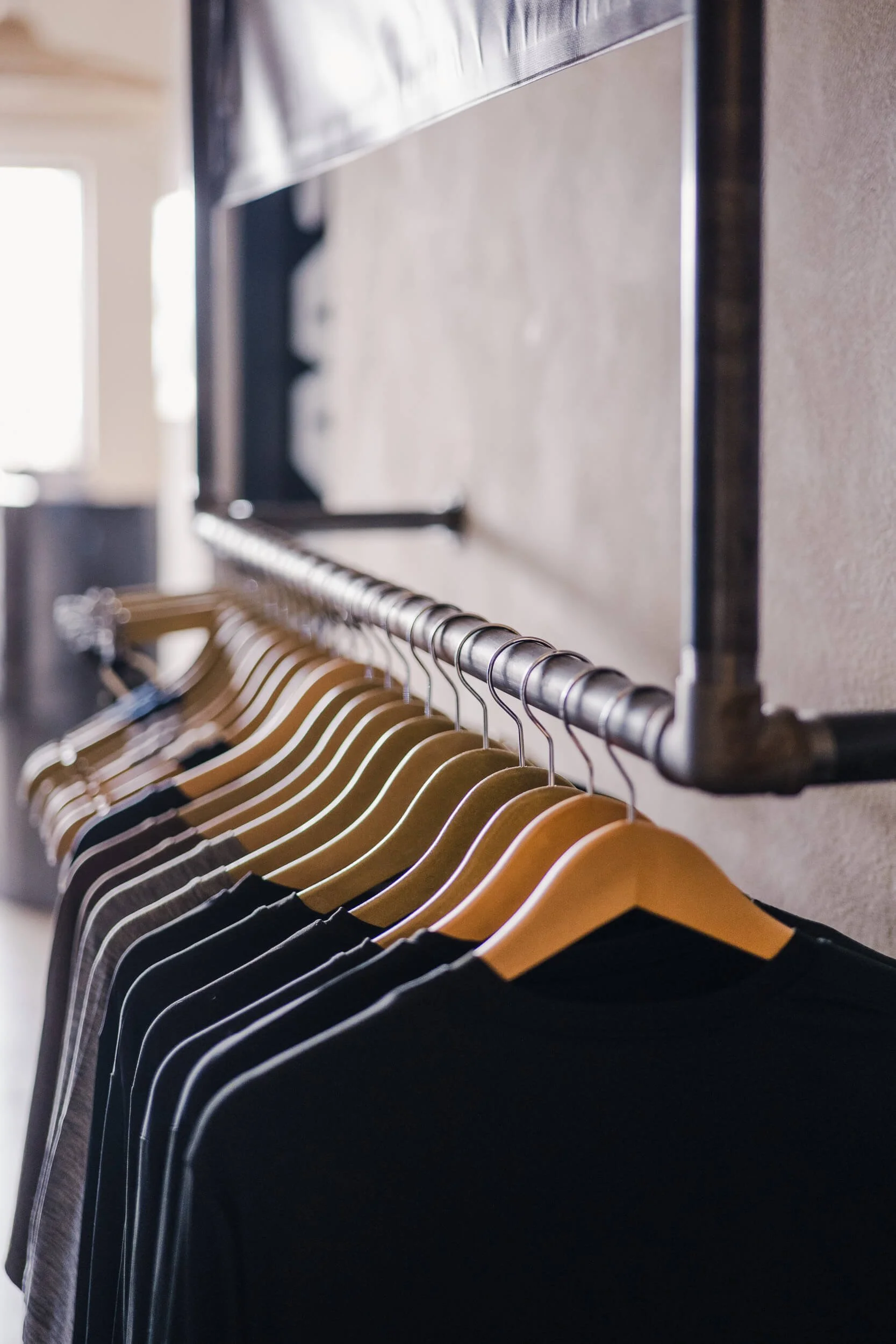 Detail of black t-shirts hanging on wooden hangers on a raw, industrial pipe clothing rack