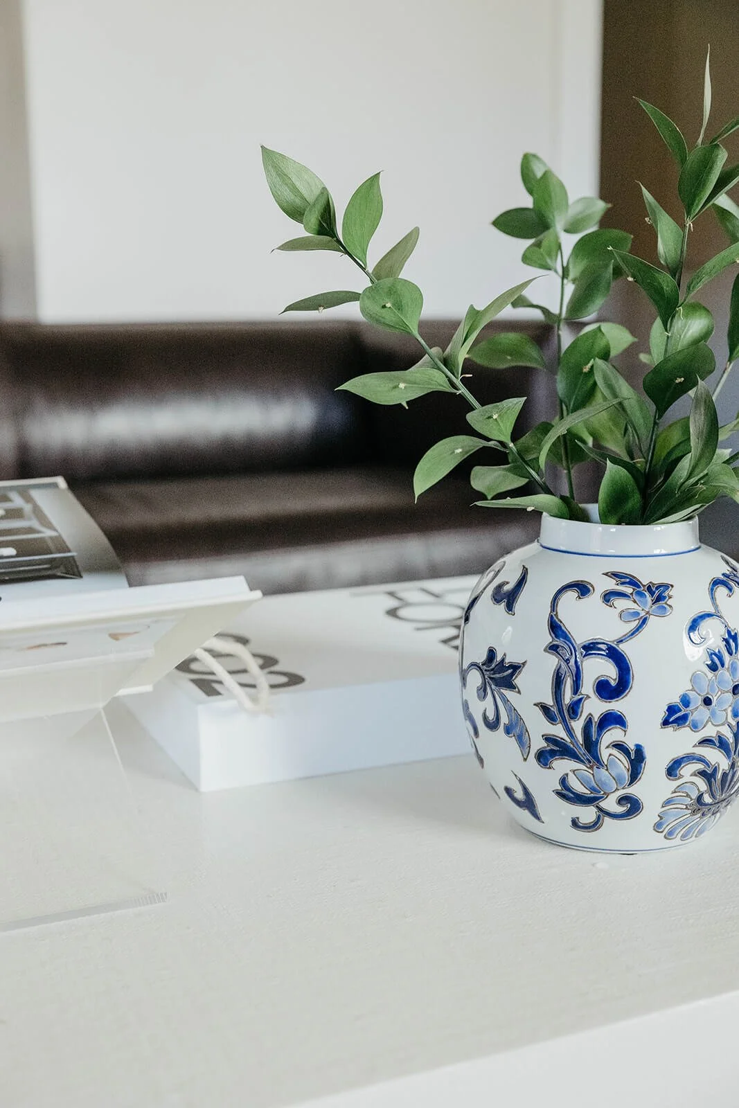 Close-up of the white coffee table styled with a blue and white chinoiserie vase holding fresh greenery, with the dark leather sofa blurred in the background.