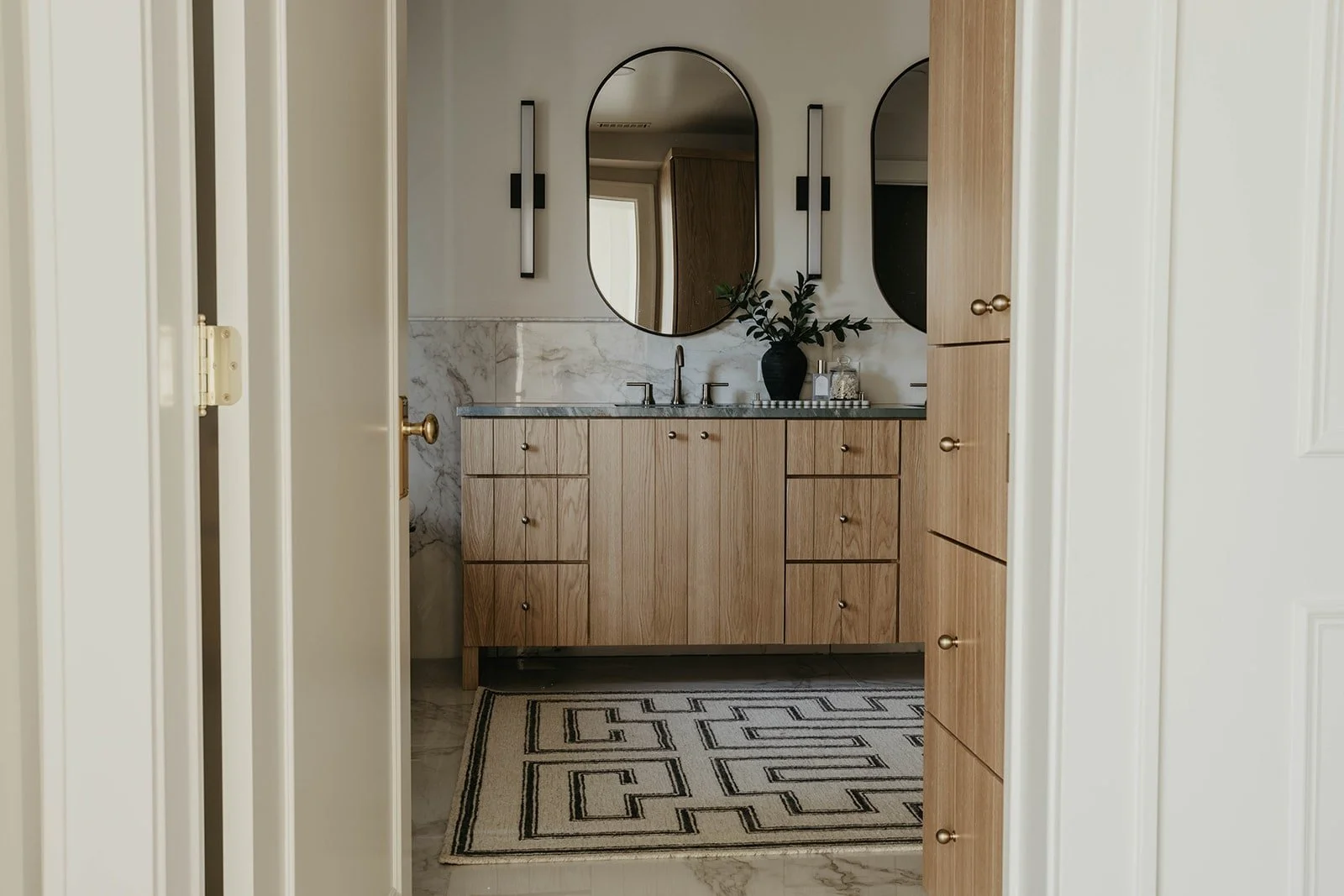 Bright, modern bathroom featuring light natural wood cabinetry, a marble countertop, and a patterned floor tile rug. Two arched mirrors hang above the vanity.
