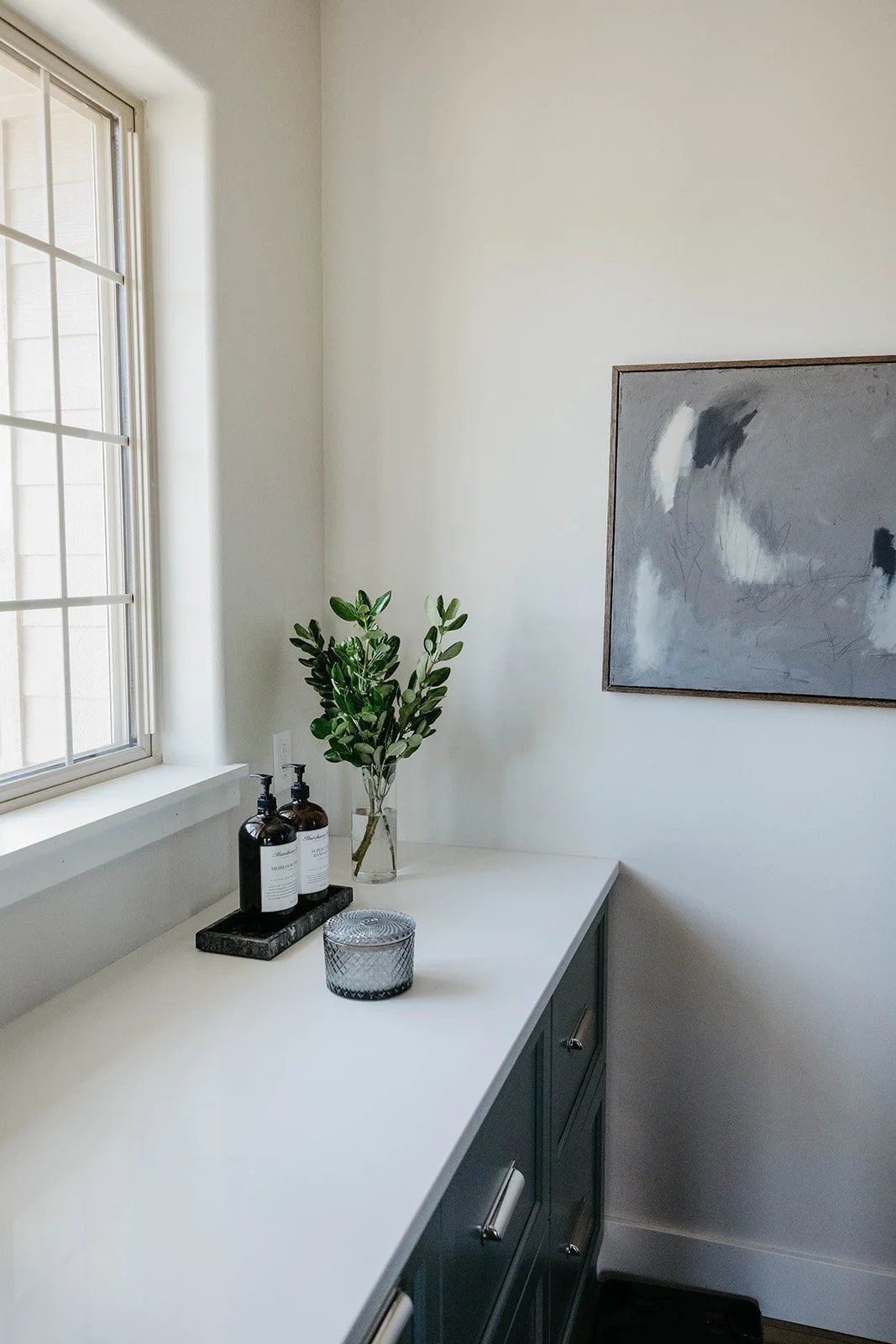 Laundry room countertop close-up with a large window and dark gray-green cabinetry. The counter is styled with soap and a framed abstract painting.