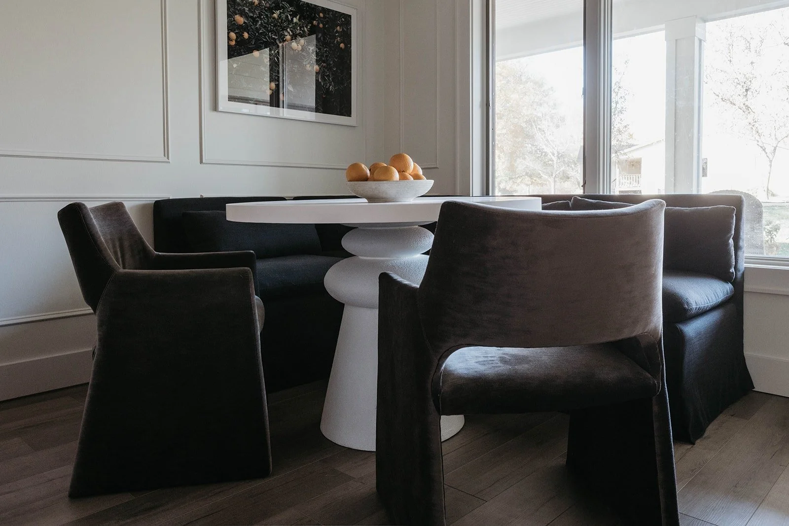 Wide view of the breakfast nook with a white round pedestal table, dark gray upholstered chairs, and a built-in banquette, positioned in front of a large window.