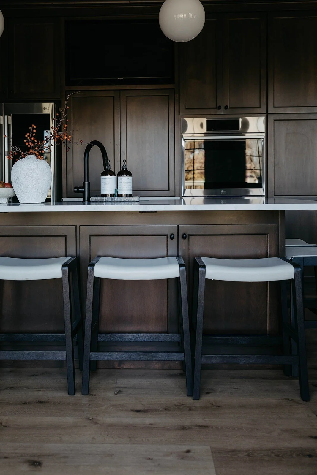 Close-up of three gray and black counter stools tucked under the kitchen island, with the built-in ovens visible in the dark cabinetry behind them.