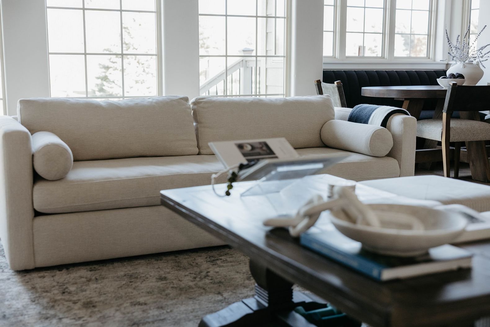 Close-up of a deep, neutral sectional sofa and a dark wood coffee table styled with decorative plates, with large windows providing ample light.