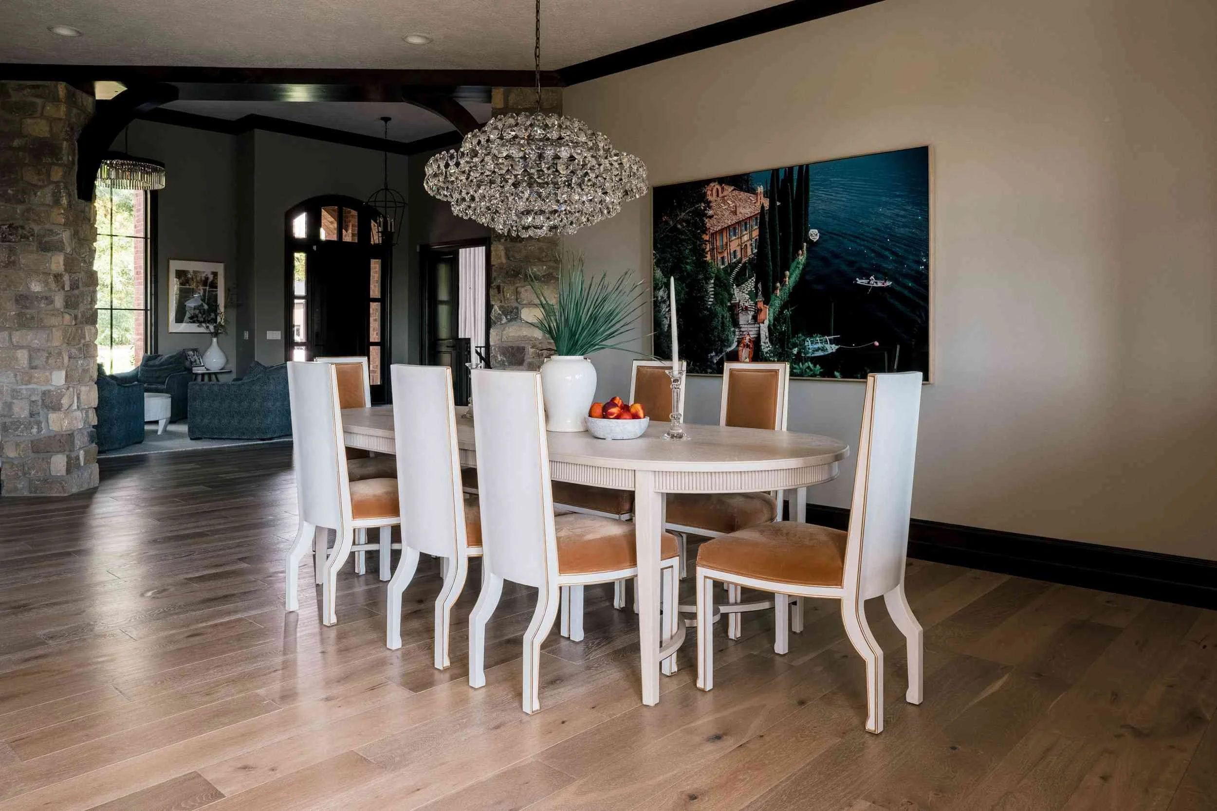 Elegant oval light-wood dining table with white and tan chairs, illuminated by a large crystal chandelier, featuring a colorful, modern piece of art on the wall.