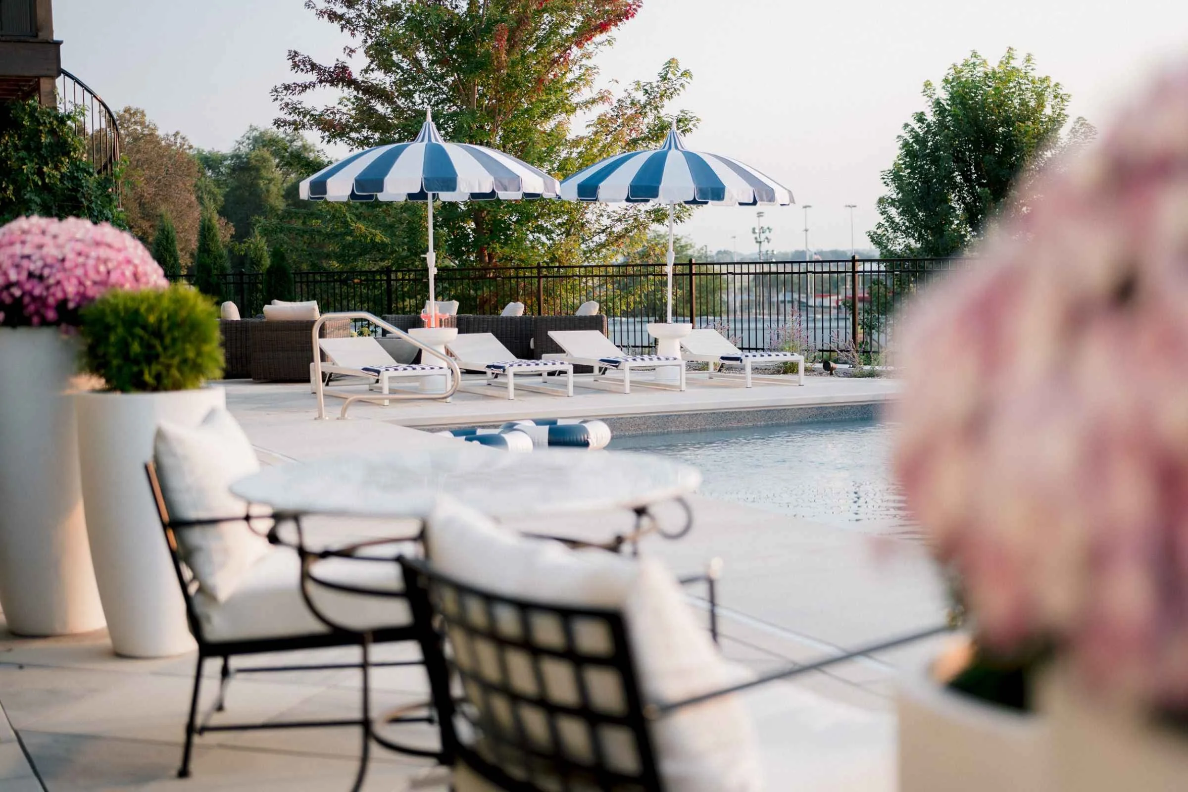 Poolside seating area with dark woven armchairs with white cushions and a distant view of the pool and the surrounding landscape, framed by a white planter of pink flowers in the foreground.