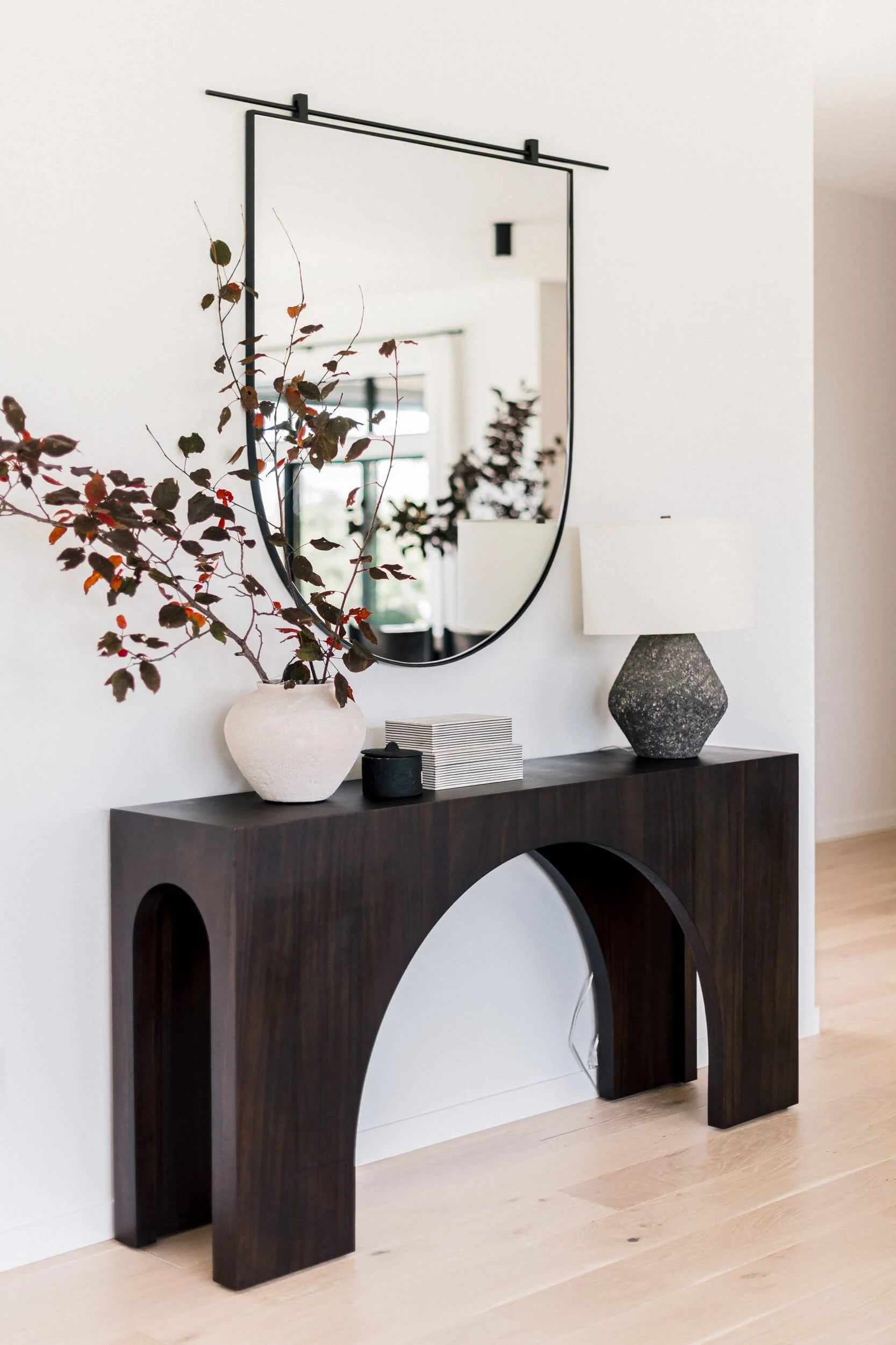 Modern entryway featuring a dark arched wood console table, a large rounded black-framed mirror, and styled with a lamp and vase with deep red foliage.
