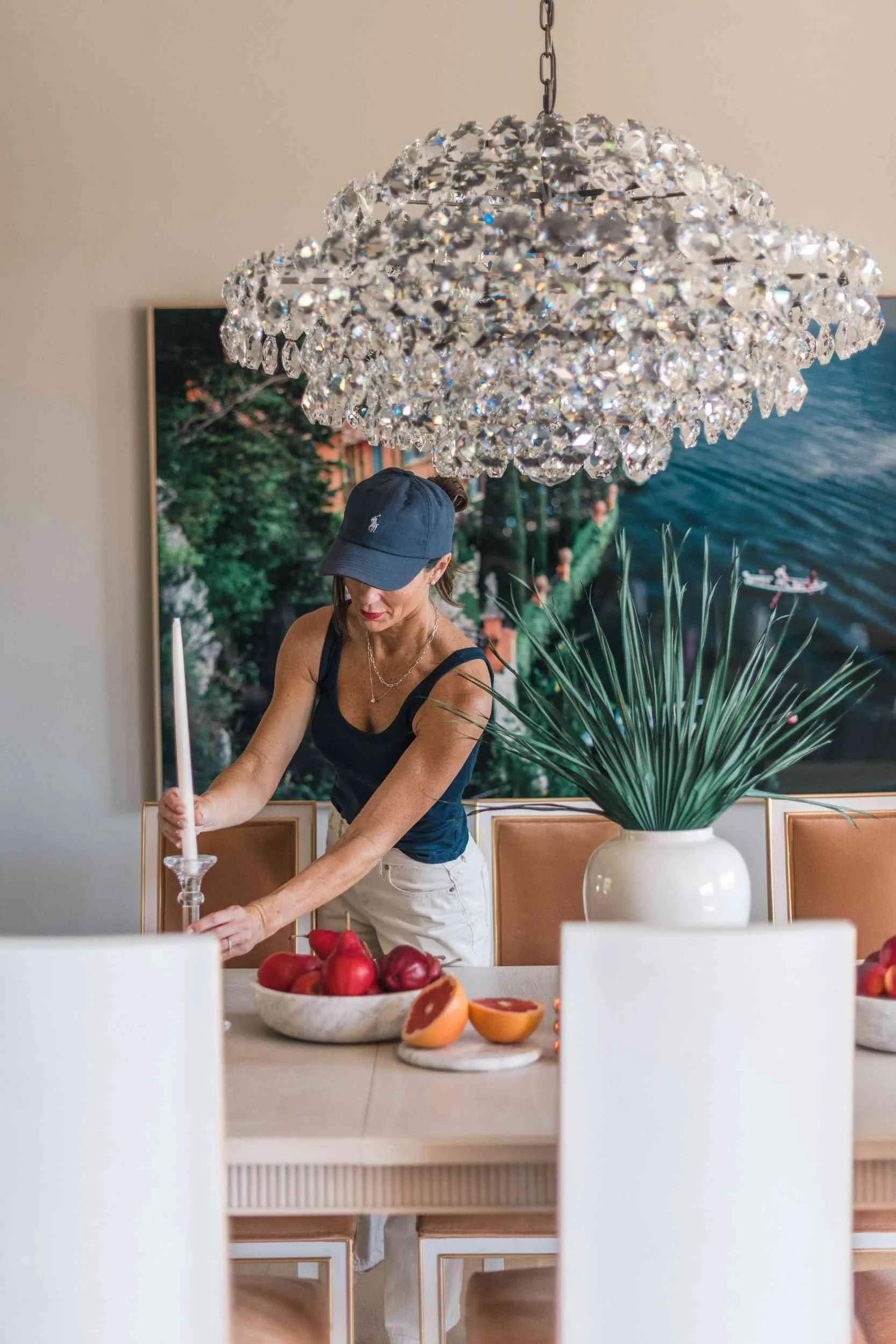 Christy Steen styling a dining table, adjusting a white candle holder next to a bowl of fruit under a large, sparkling crystal chandelier.