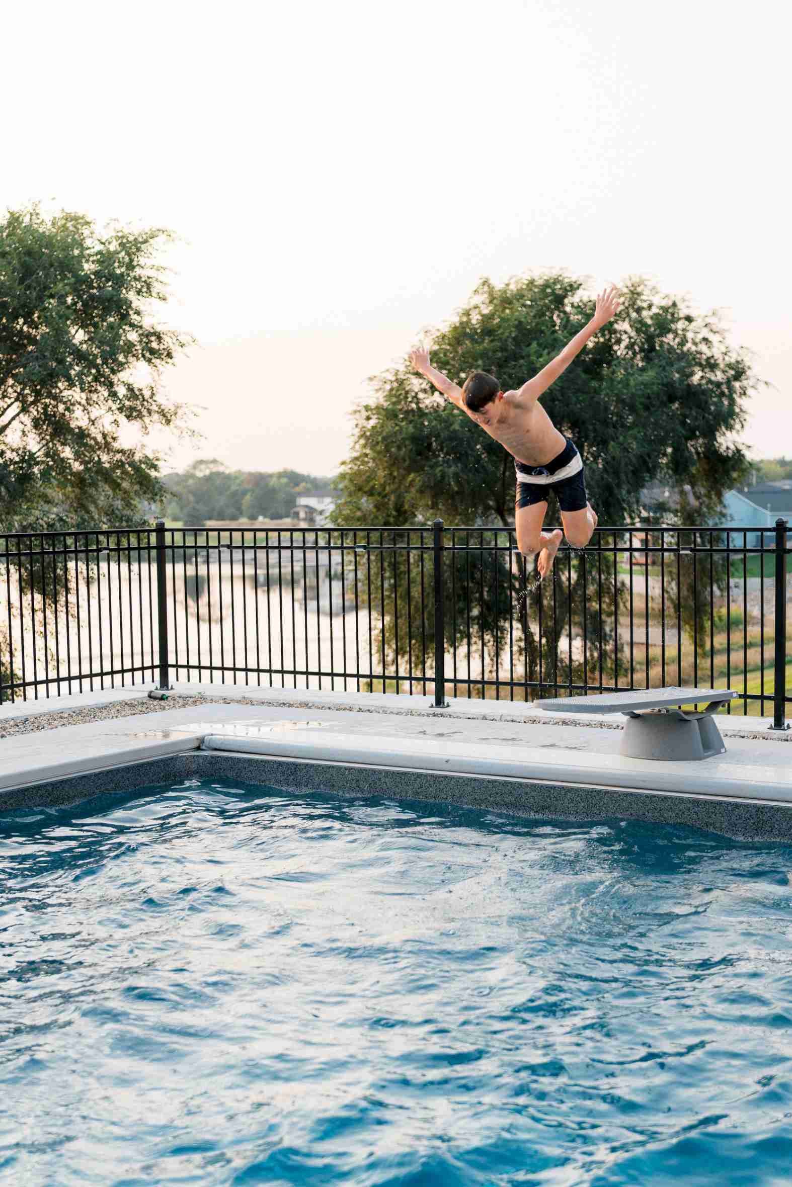 Boy jumping off a diving board into the pool against an expansive evening sky, with the black metal fence and landscape visible.