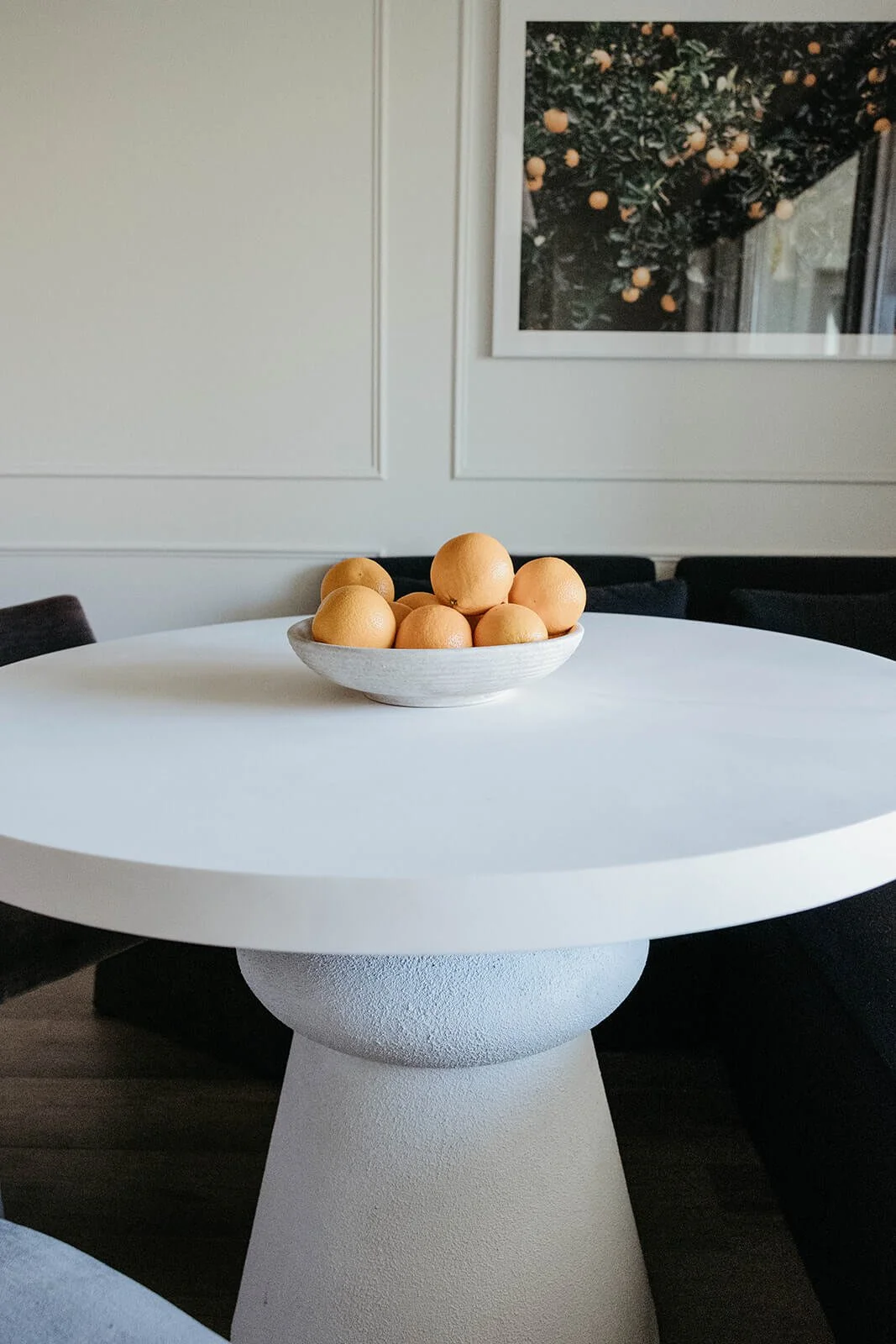 Close-up of the white round dining table with a heavy, textured pedestal base and a bowl of oranges centered on the top.
