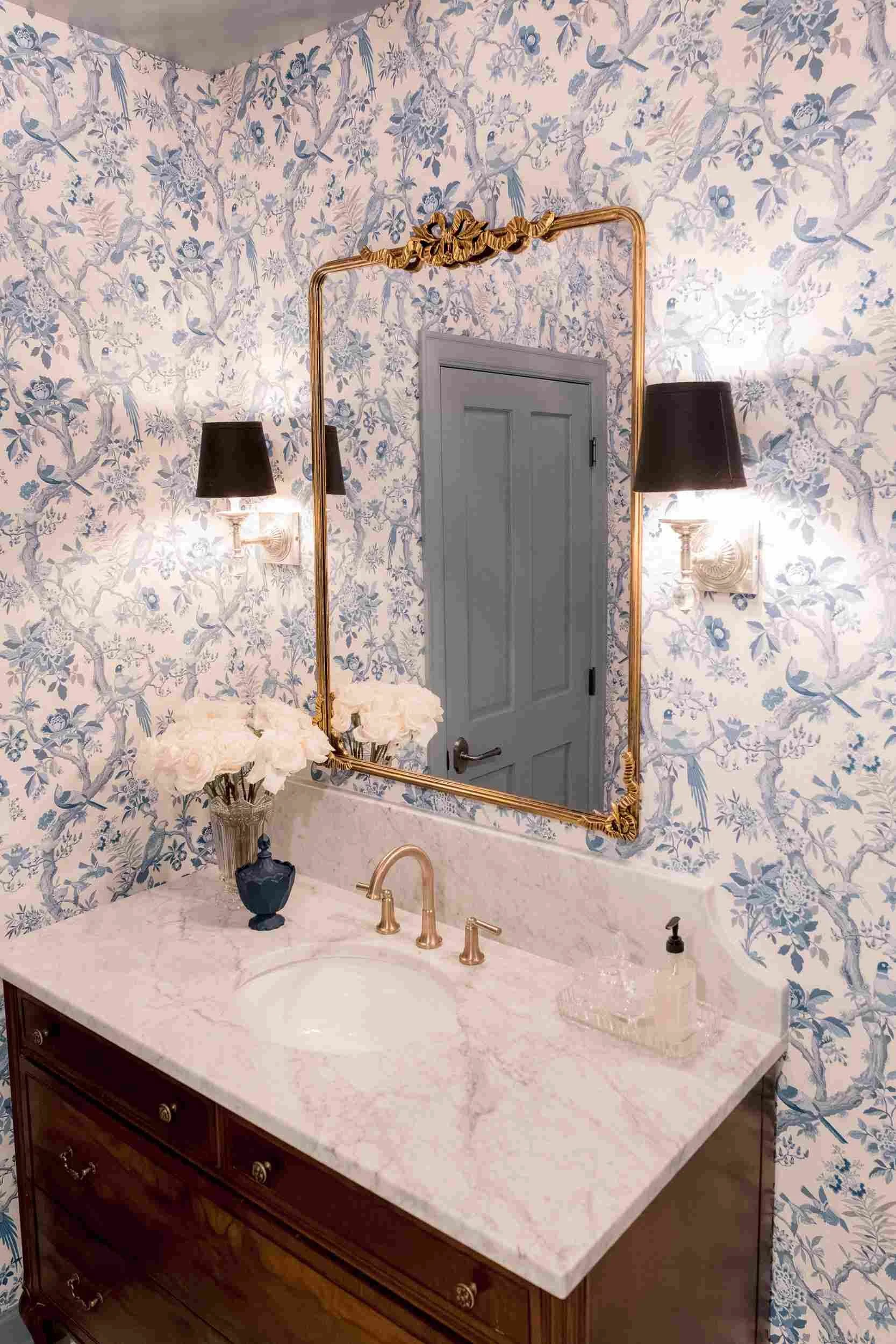 View of the powder room vanity with blue toile wallpaper, a gilt mirror, and a large bouquet of cream-colored roses on the marble countertop.