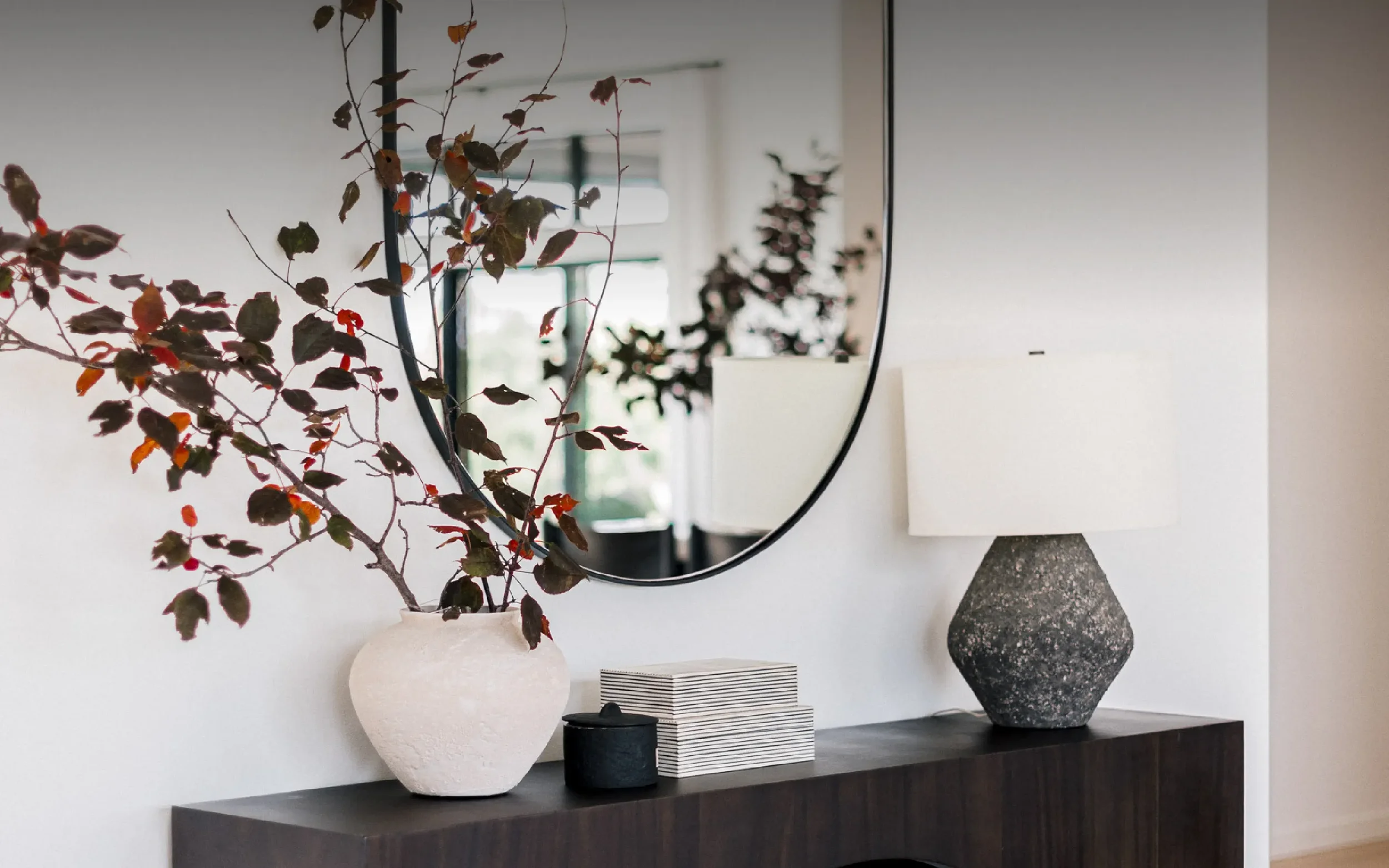Modern entryway console table with a dark arched wood base and a black metal frame oval mirror hanging above. The table is styled with a textured gray lamp, a white vase holding deep red branches, and decorative boxes.