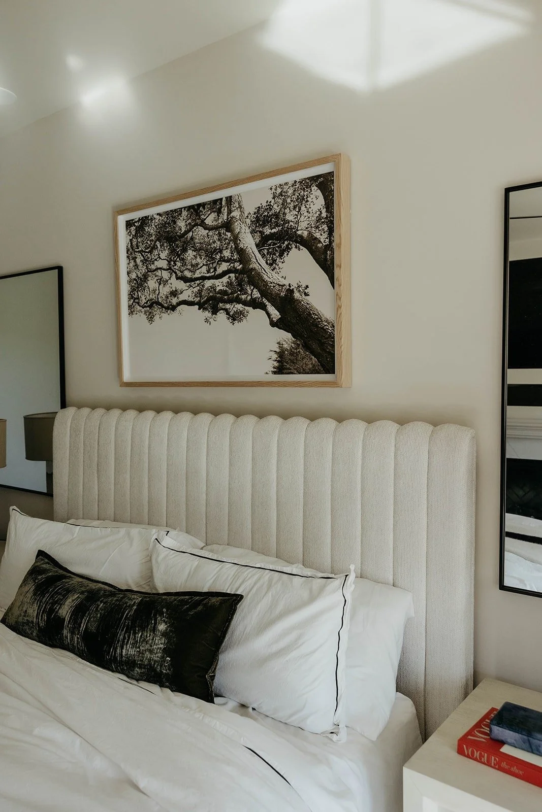Close-up of the master bed with a channel-tufted beige headboard, layered white bedding, and a black velvet accent pillow. A wooden framed tree print hangs above.