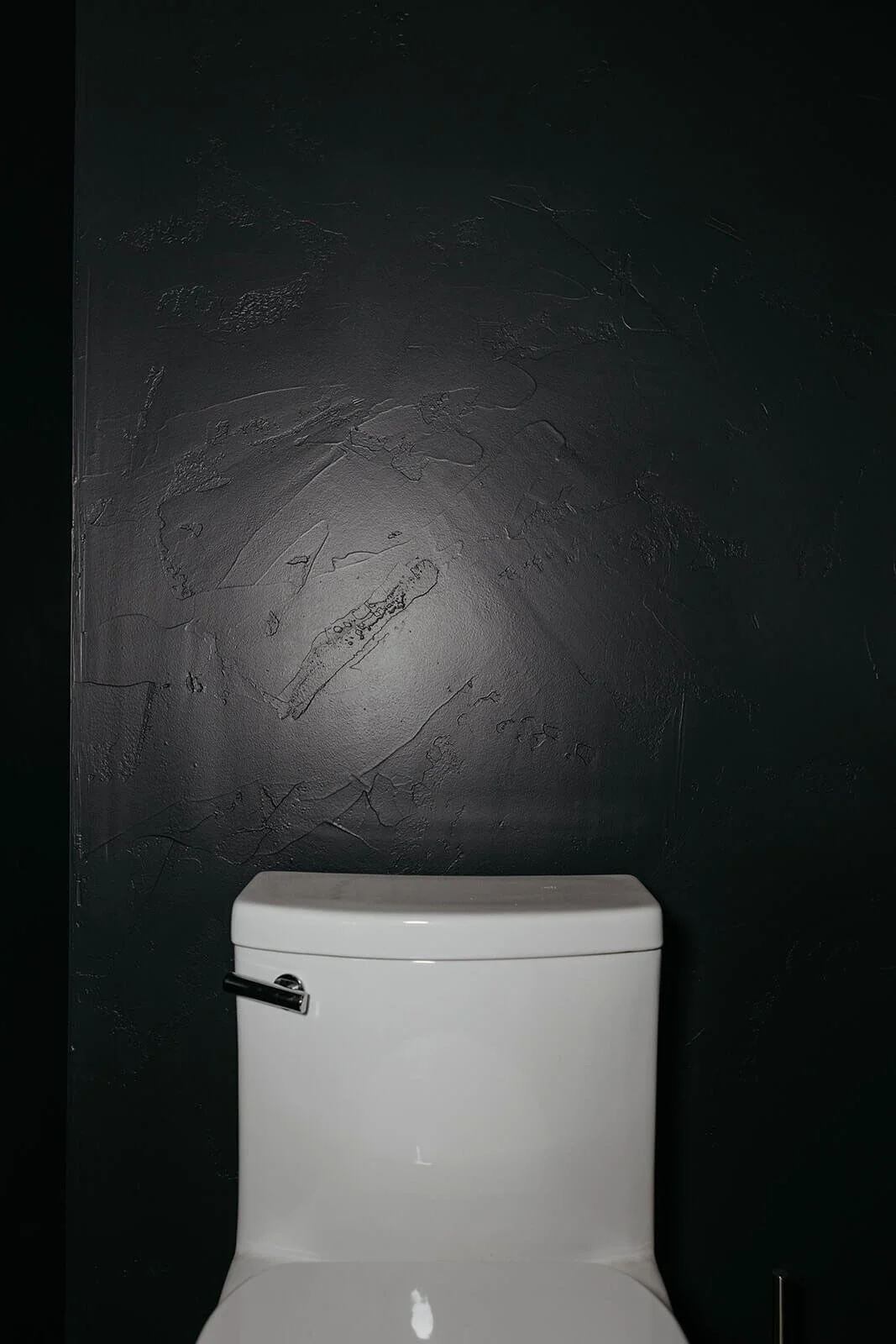 Close-up of the white toilet tank set against the heavily textured dark charcoal or black wall surface in the powder room, highlighting the distressed finish. 
