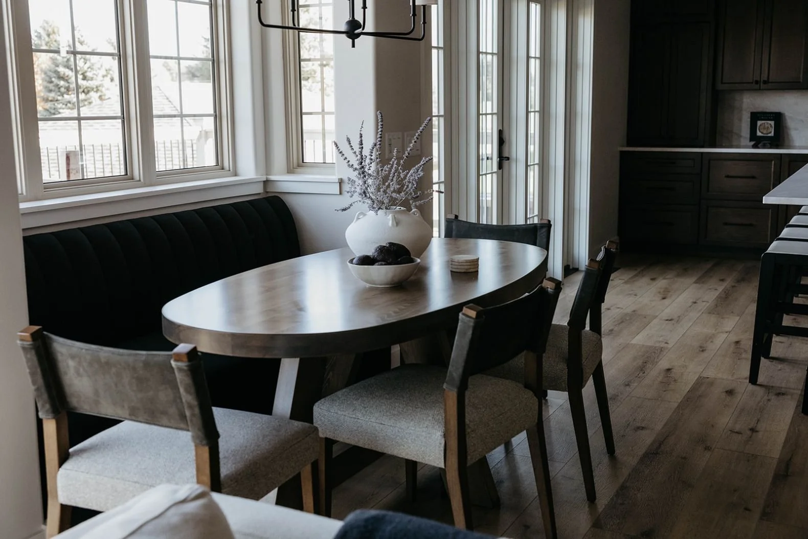 Wide view of the dark dining banquette nook with the round wood table, highlighting the natural light from the windows.