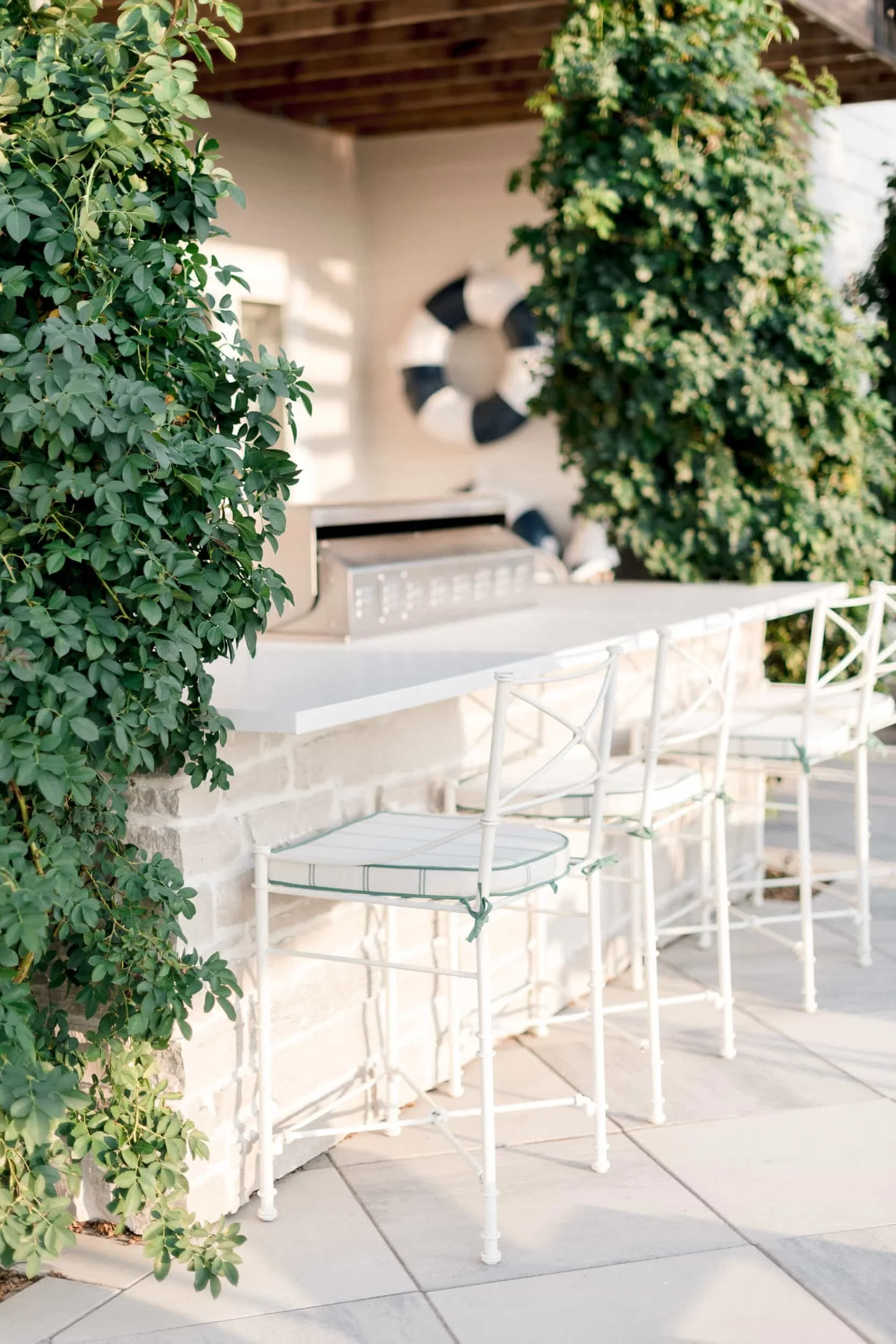 White outdoor bar area with three metal barstools that have light blue and white striped cushions. The space features a white counter and is surrounded by tall, vibrant greenery.