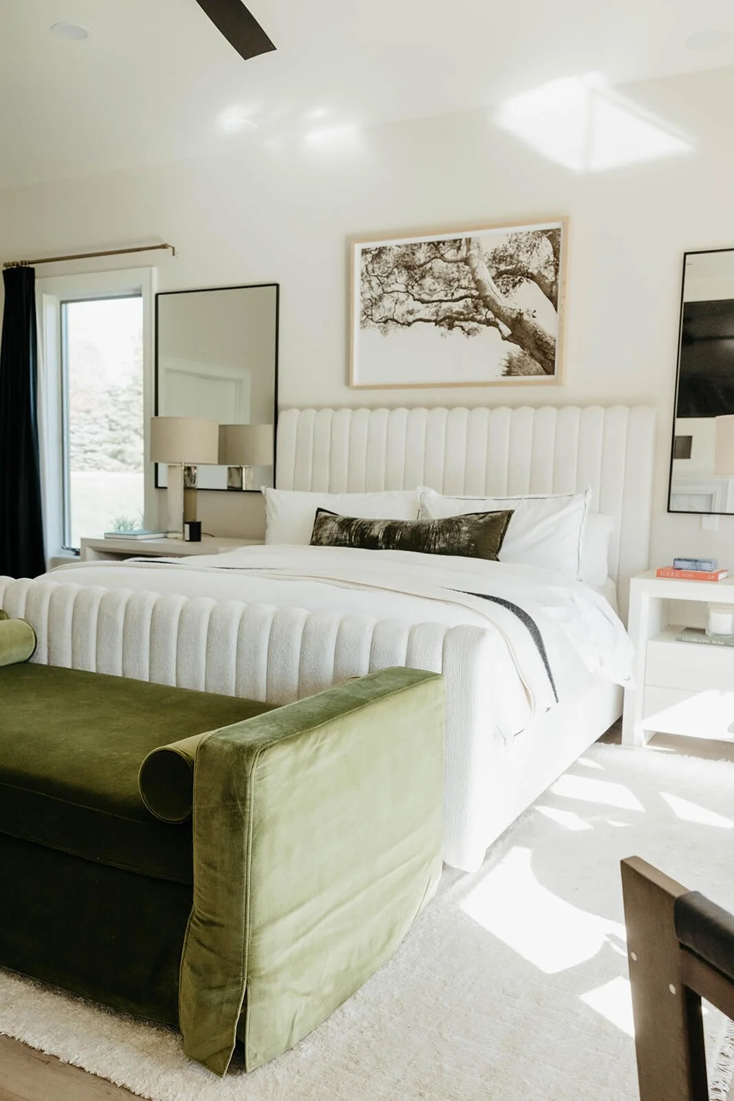 Bright master bedroom featuring a large white bed with a tufted headboard, an olive green velvet bench at the foot of the bed, and a mirrored nightstand.