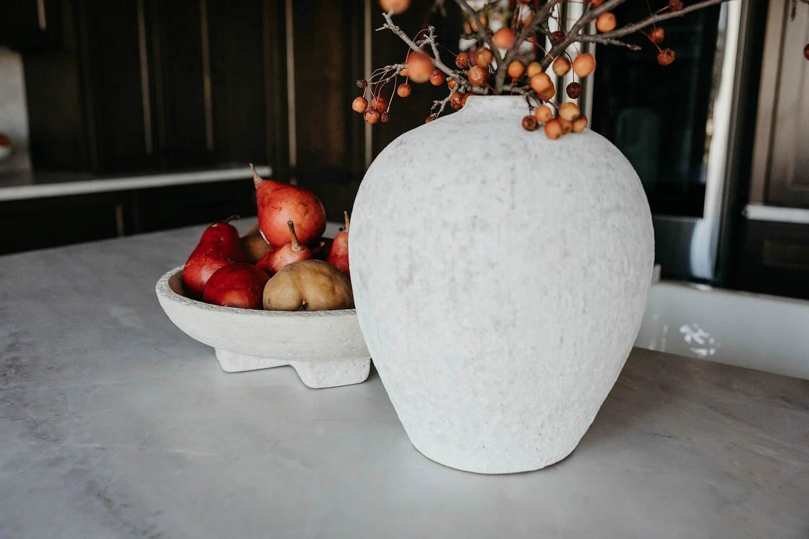 Close-up of kitchen countertop styling featuring a round stone bowl of apples and pomegranates next to a textured white vase with berries