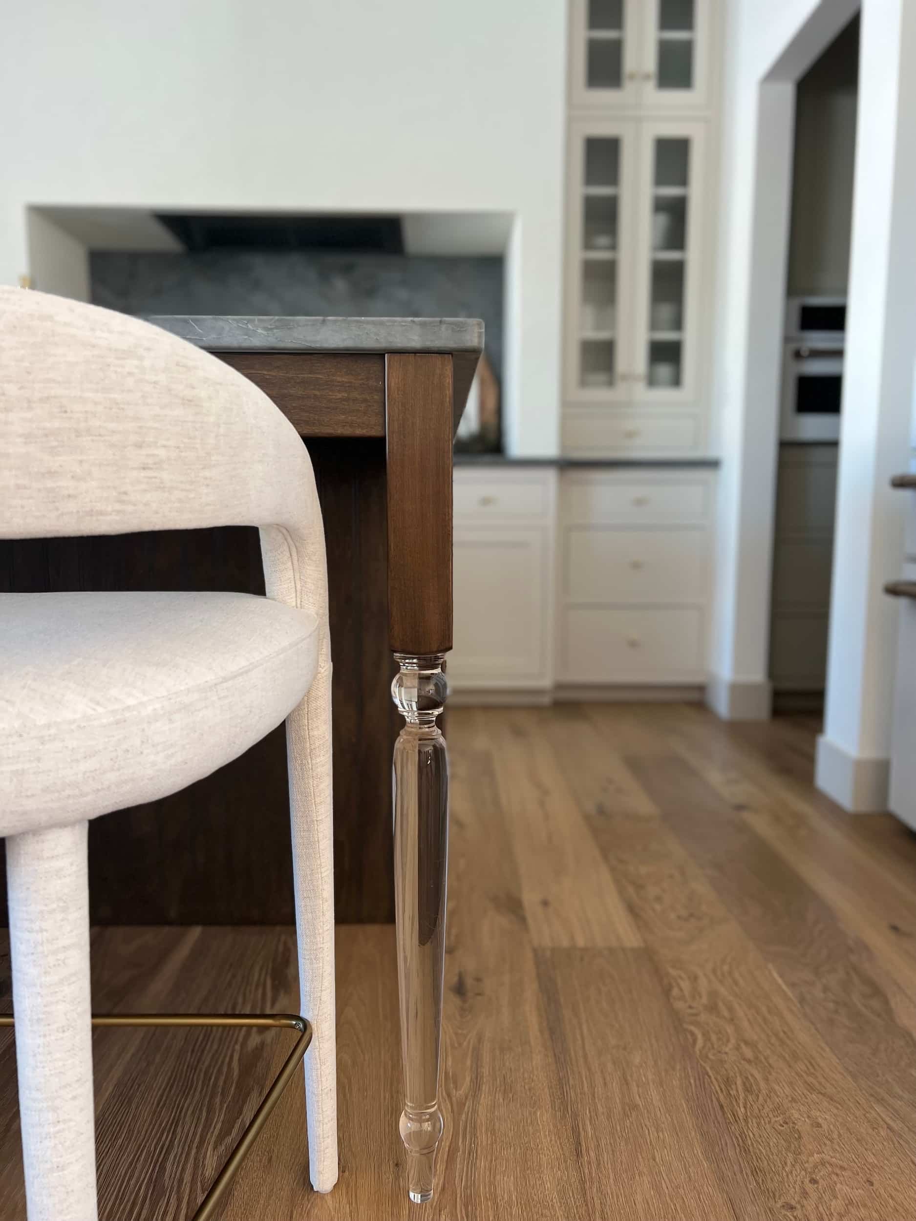 Close-up of a neutral upholstered bar stool next to the kitchen island, highlighting the dark wood and clear acrylic island leg detail.