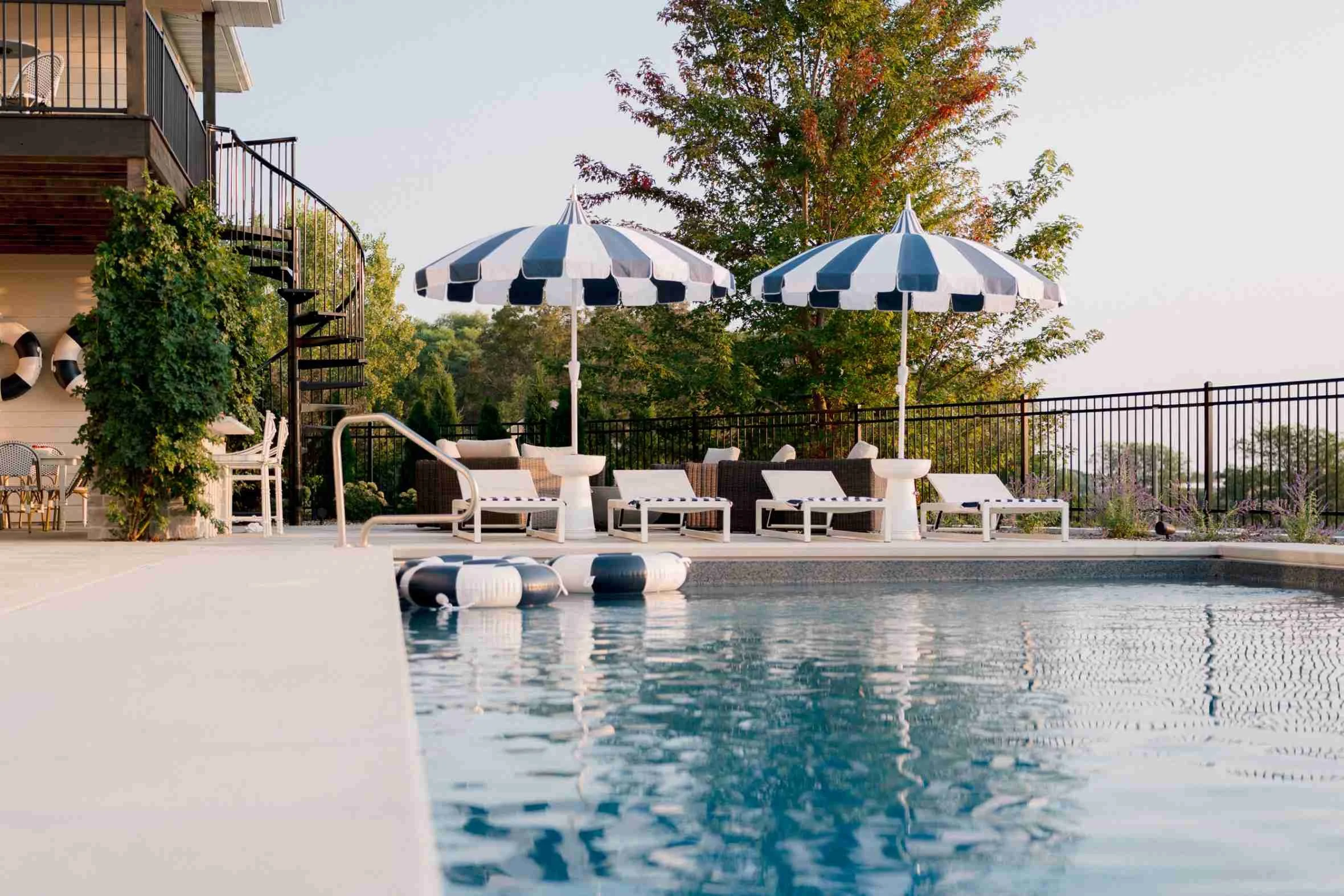 Wide view of the pool deck showing multiple white chaise lounges, striped umbrellas, and a black spiral staircase leading down from the house exterior.