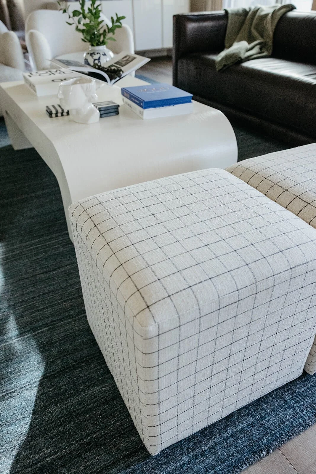 Detail shot of a contemporary living room showing a white plaid upholstered ottoman next to the sculpted white coffee table and dark leather sofa, all resting on a textured blue rug.